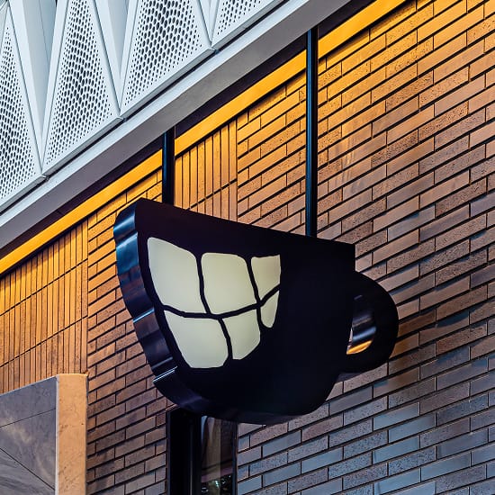 Illuminated black coffee cup shaped projecting blade sign with smile logo mounted on brick exterior at Virgin Hotels Dallas - custom dimensional blade signage and experiential retail branding design by RSM Design, a leading architectural signage and wayfinding design firm