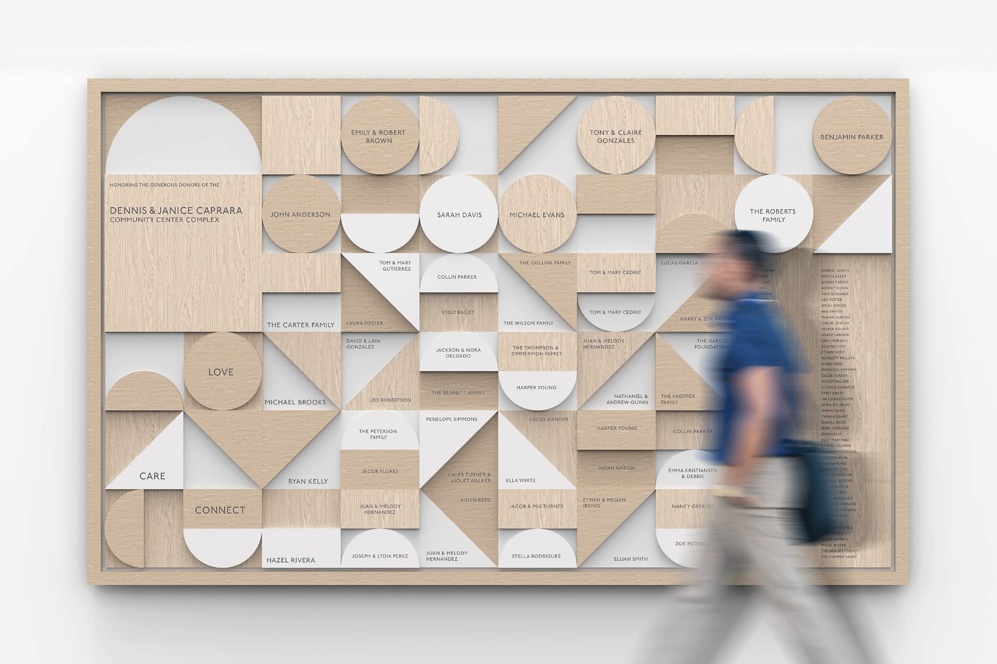 Donor recognition wall for the Dennis & Janice Caprara Community Center Complex, made of geometric wood and white circular, triangular, and rectangular tiles engraved with individual donor names and the words Love, Care, and Connect, as a man walks past.