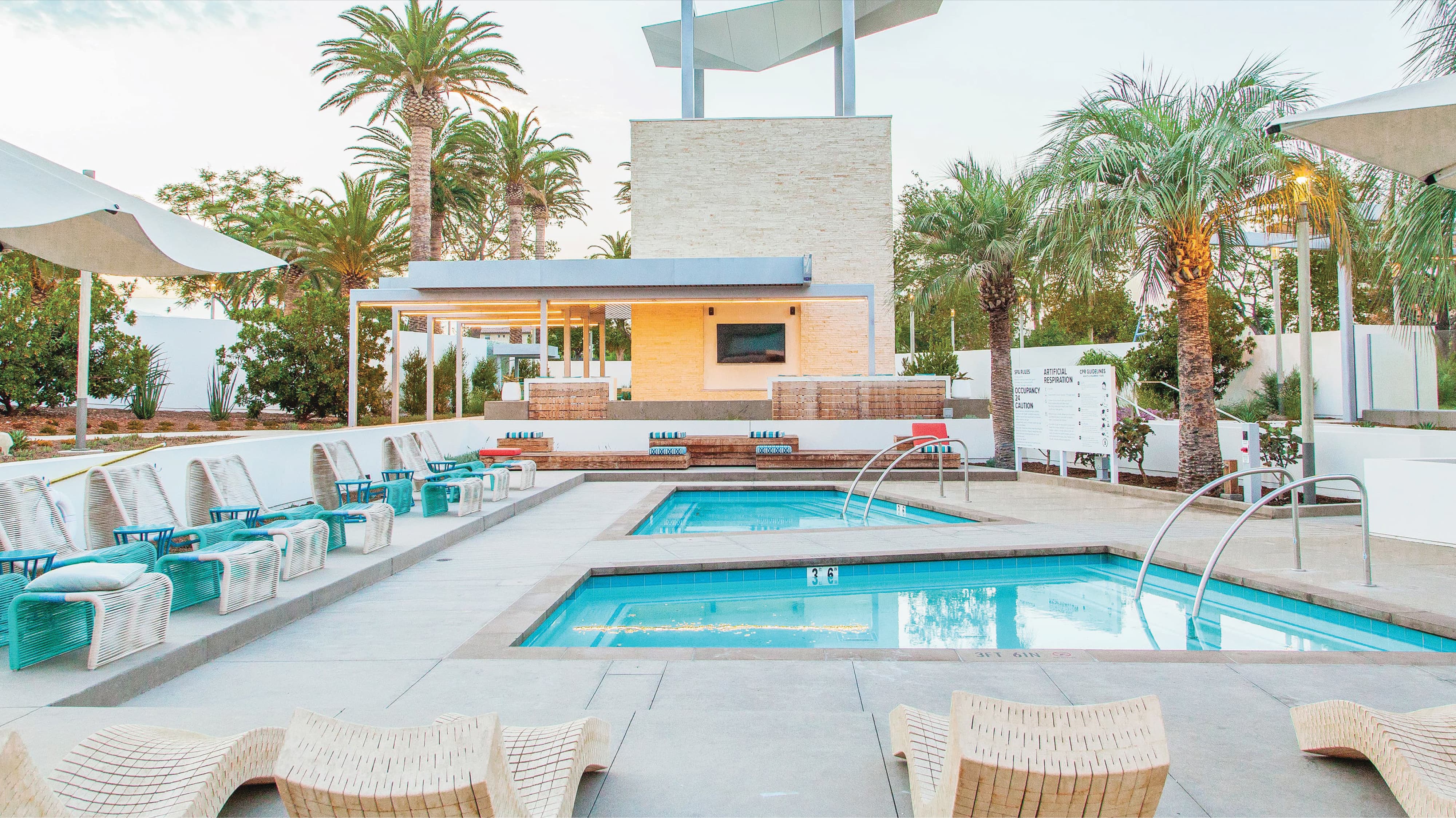 Community pool area with two rectangular pools flanked by teal lounge chairs and sculptural cream loungers, framed by palm trees, a stone-clad tower with an angular sculpture on top, and a shaded pergola seating area in the background.