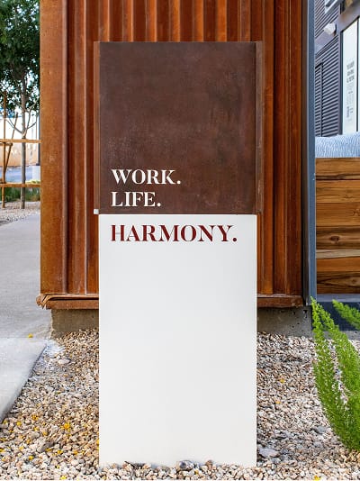 A freestanding two-toned sign at Uncommons reads "Work. Life. Harmony." with the first two lines in white text on a Corten steel upper panel and "Harmony." in crimson on a white lower panel. The sign is set in a gravel landscape bed with a succulent plant, in front of a warm wood and Corten steel building facade.