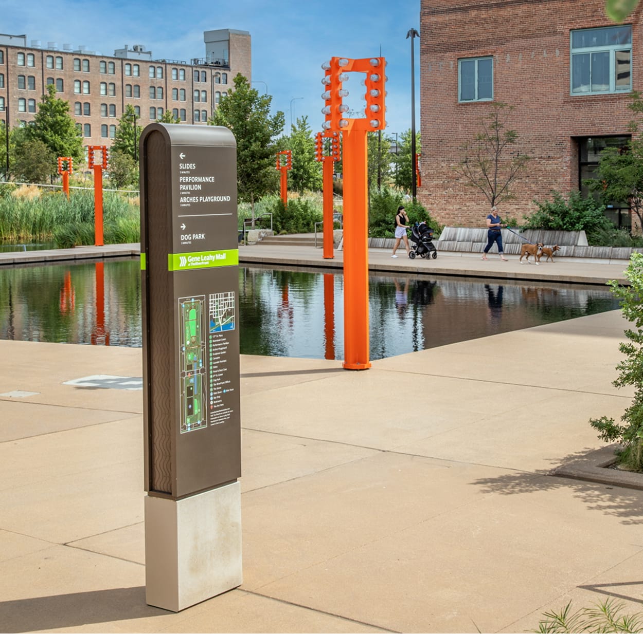 Gene Leahy Mall wayfinding kiosk at the RiverFront in downtown Omaha, Nebraska, directing visitors to the Slides, Performance Pavilion, Arches Playground, and Dog Park, with orange light tower sculptures reflecting in the canal and pedestrians strolling in the background