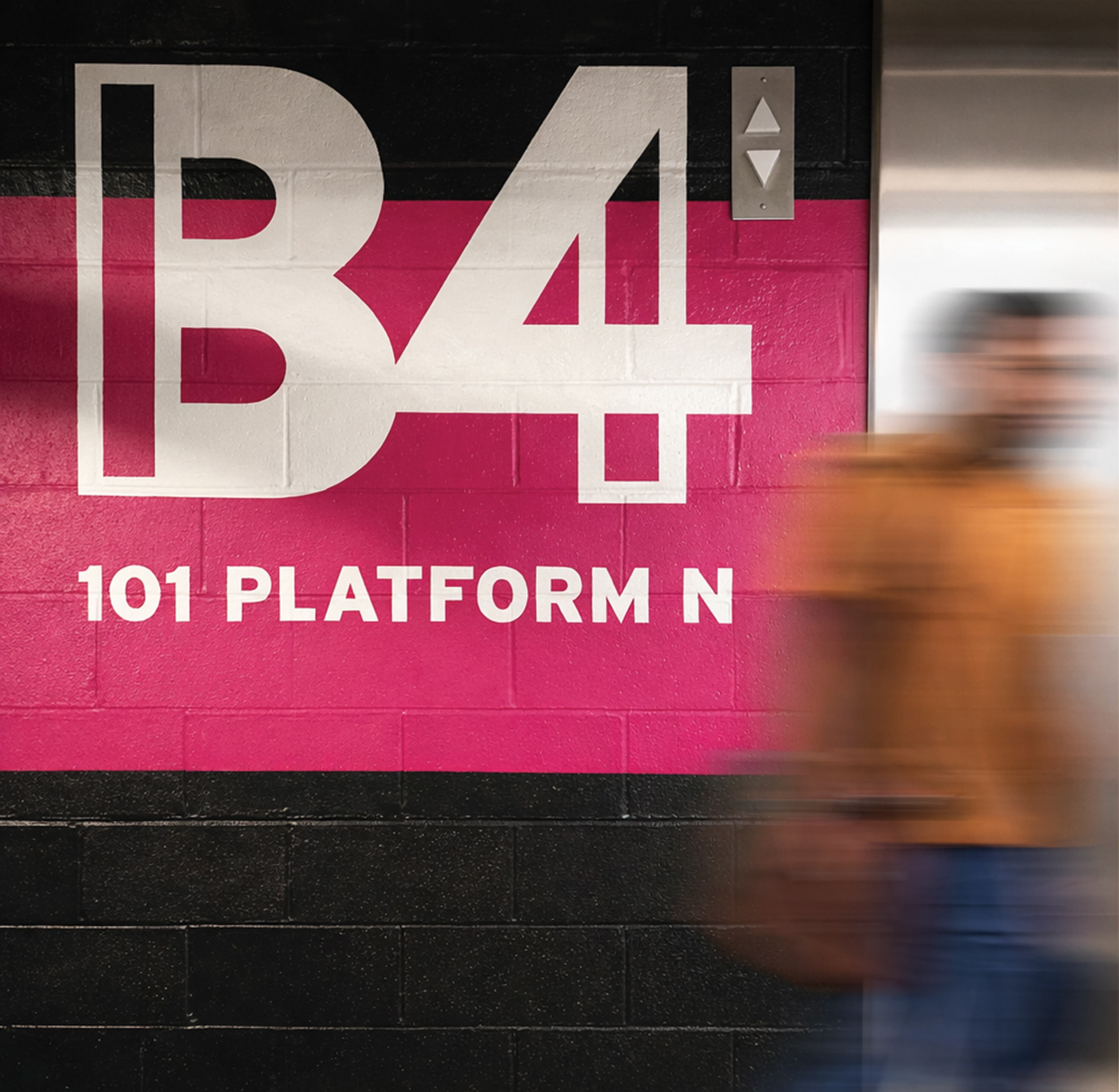 Bold "B4 – 101 Platform N" level identification painted directly on a concrete block wall in white and magenta as a pedestrian passes by an elevator, showcasing experiential graphics and wayfinding design for a parking garage environment by RSM Design.