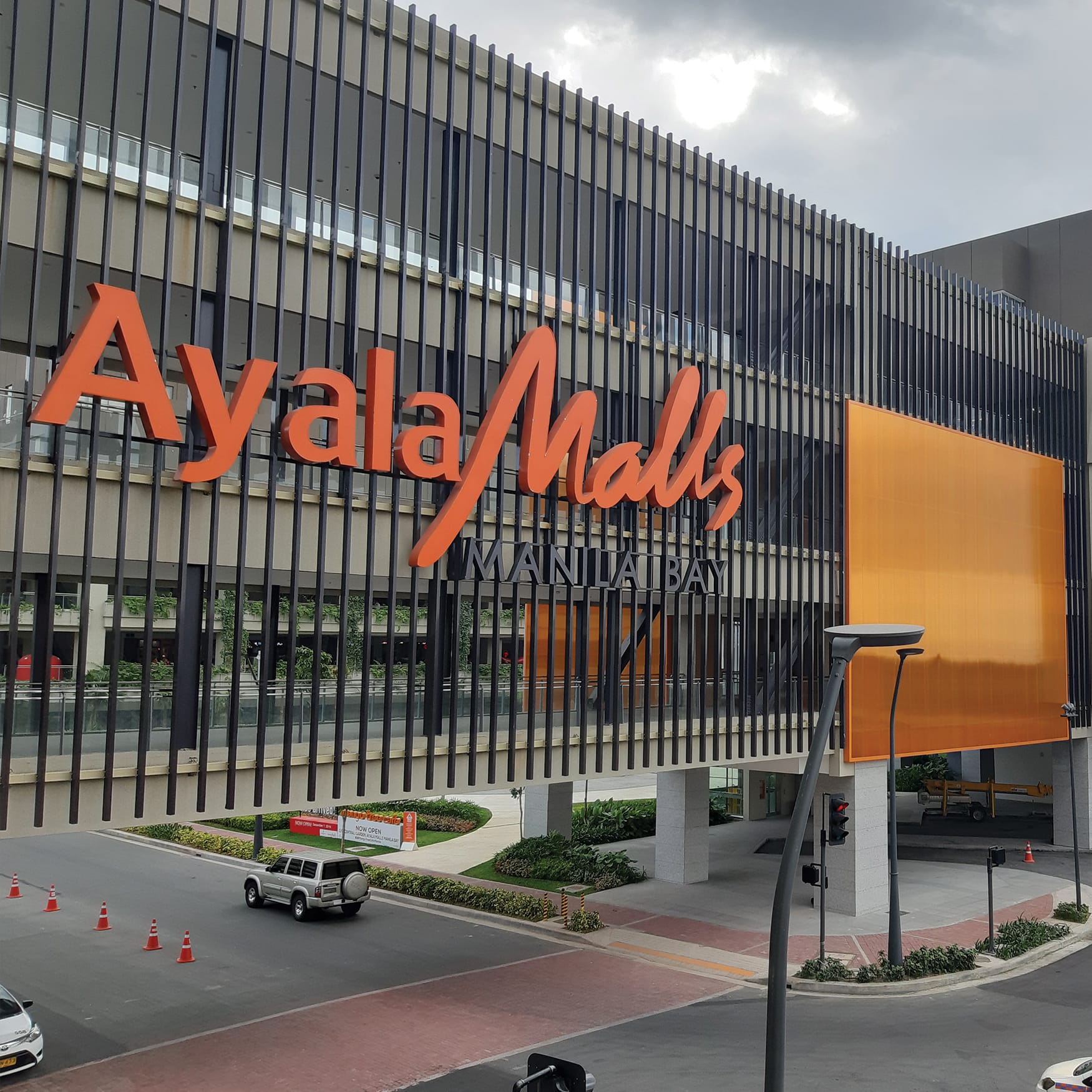 Exterior facade and overhead walkway of Ayala Malls Manila Bay in Parañaque, Philippines featuring large orange branding signage and modern slat architecture over a street.