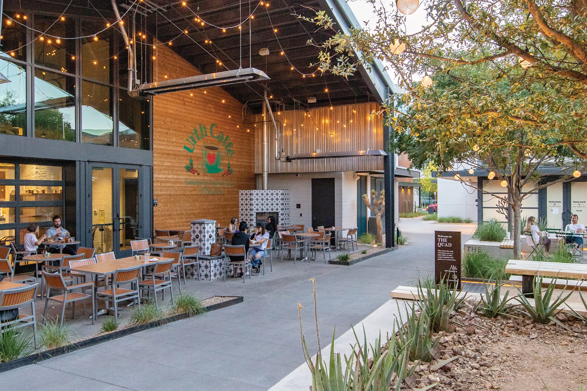 Outdoor courtyard at Uncommons at dusk, with people dining on the patio of Urth Caffé, whose logo is painted on a warm wood-clad exterior wall. String lights hang overhead, and a decorative tiled fireplace anchors the seating area. Desert plantings including agave and ornamental grasses line the walkway. The "Welcome to The Quad" sign is visible in the middle distance, with additional retail storefronts and seating areas extending down the pedestrian corridor.