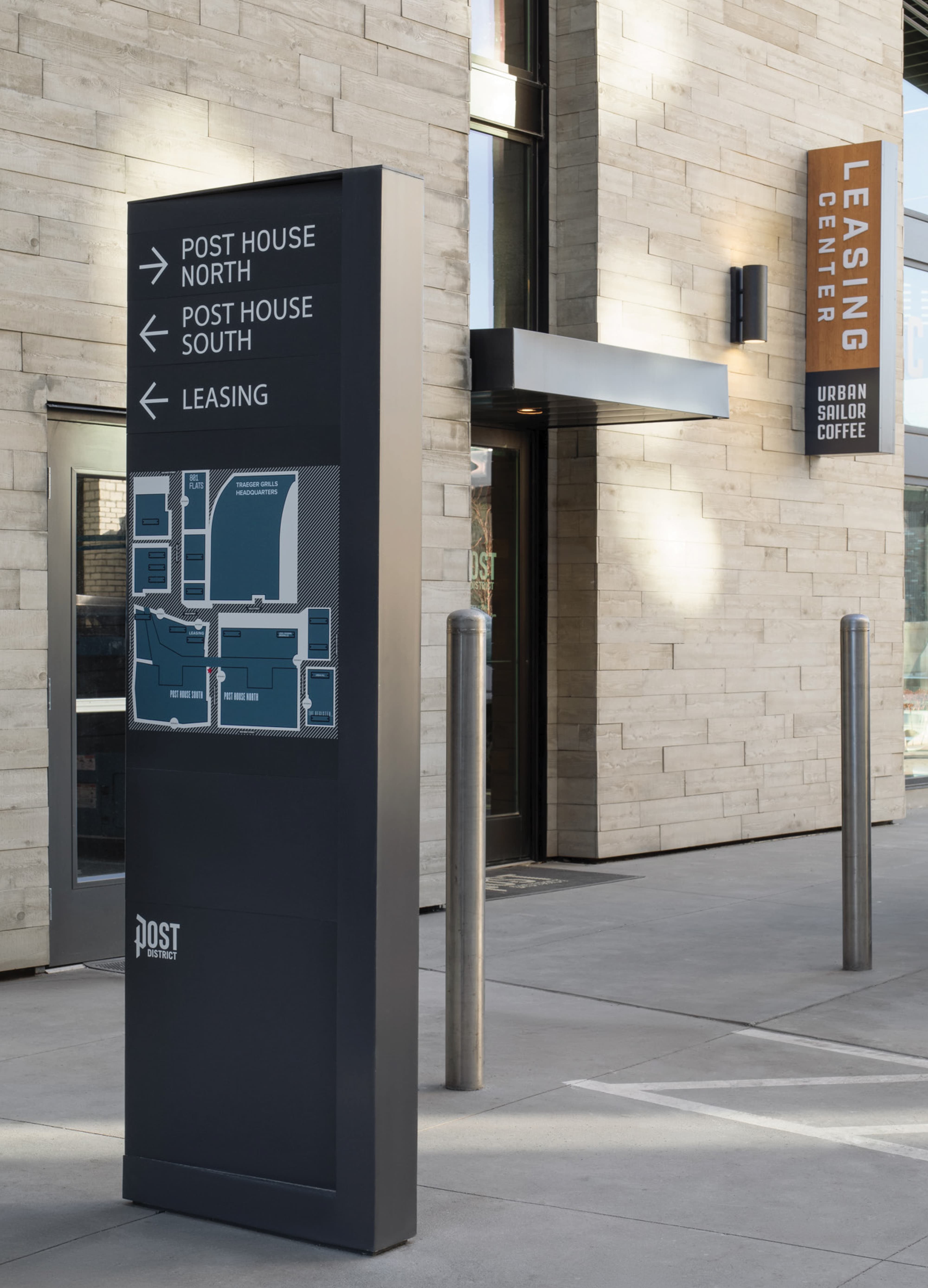 Exterior wayfinding directory pylon sign for Post District featuring directional arrows to Post House North, Post House South, and Leasing, along with an integrated site map — paired with a Leasing Center blade sign and Urban Sailor Coffee tenant identification sign on a stone building facade, showcasing RSM Design's wayfinding signage design and environmental graphic design for a mixed-use residential community.