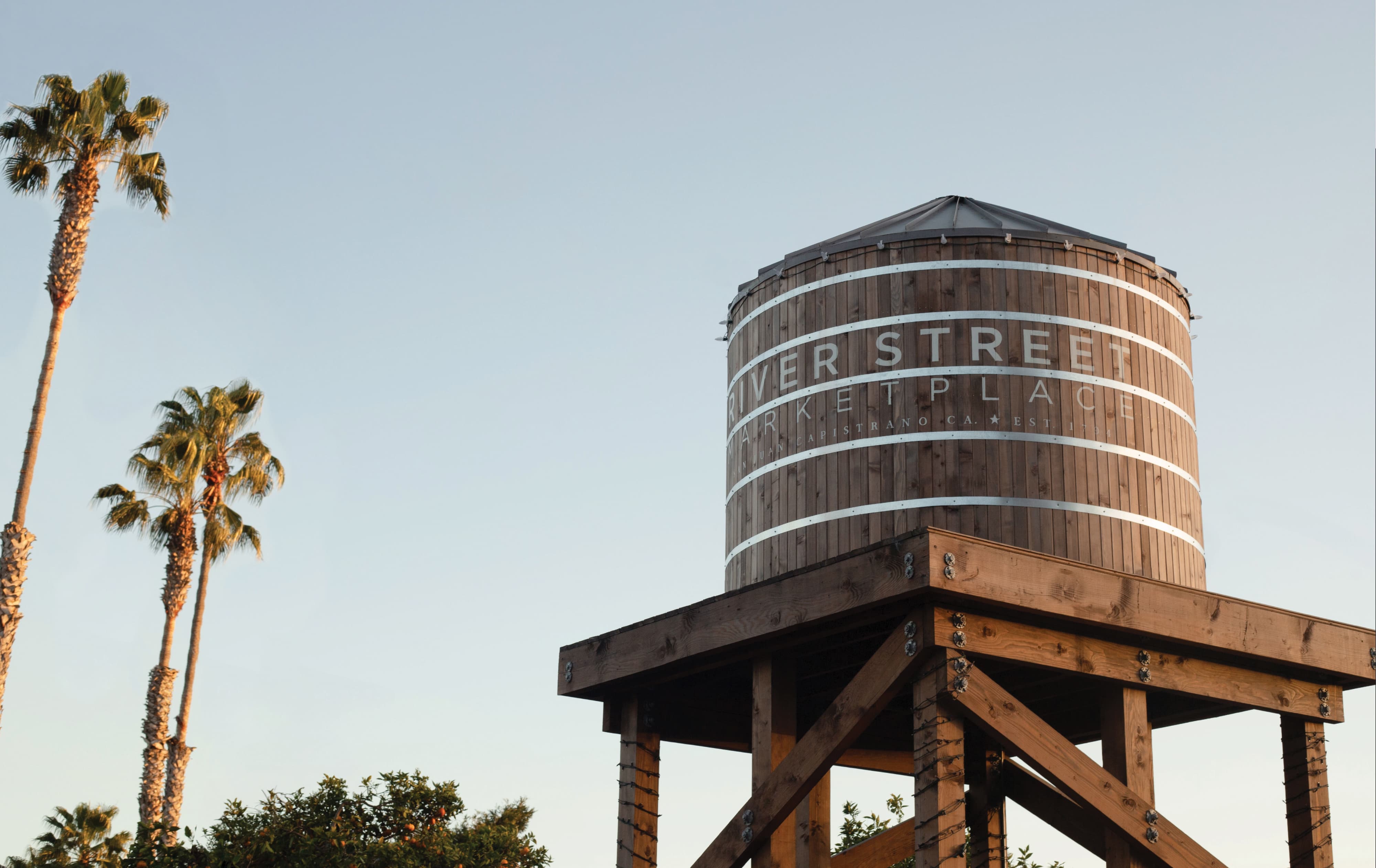 RSM Design landmark identity and placemaking design for River Street Marketplace in San Juan Capistrano, California, featuring a large-scale branded water tower with applied typography against a warm evening sky and palm trees, serving as an iconic wayfinding landmark and experiential graphic element that roots the mixed-use development in its regional identity and streetscape context.
