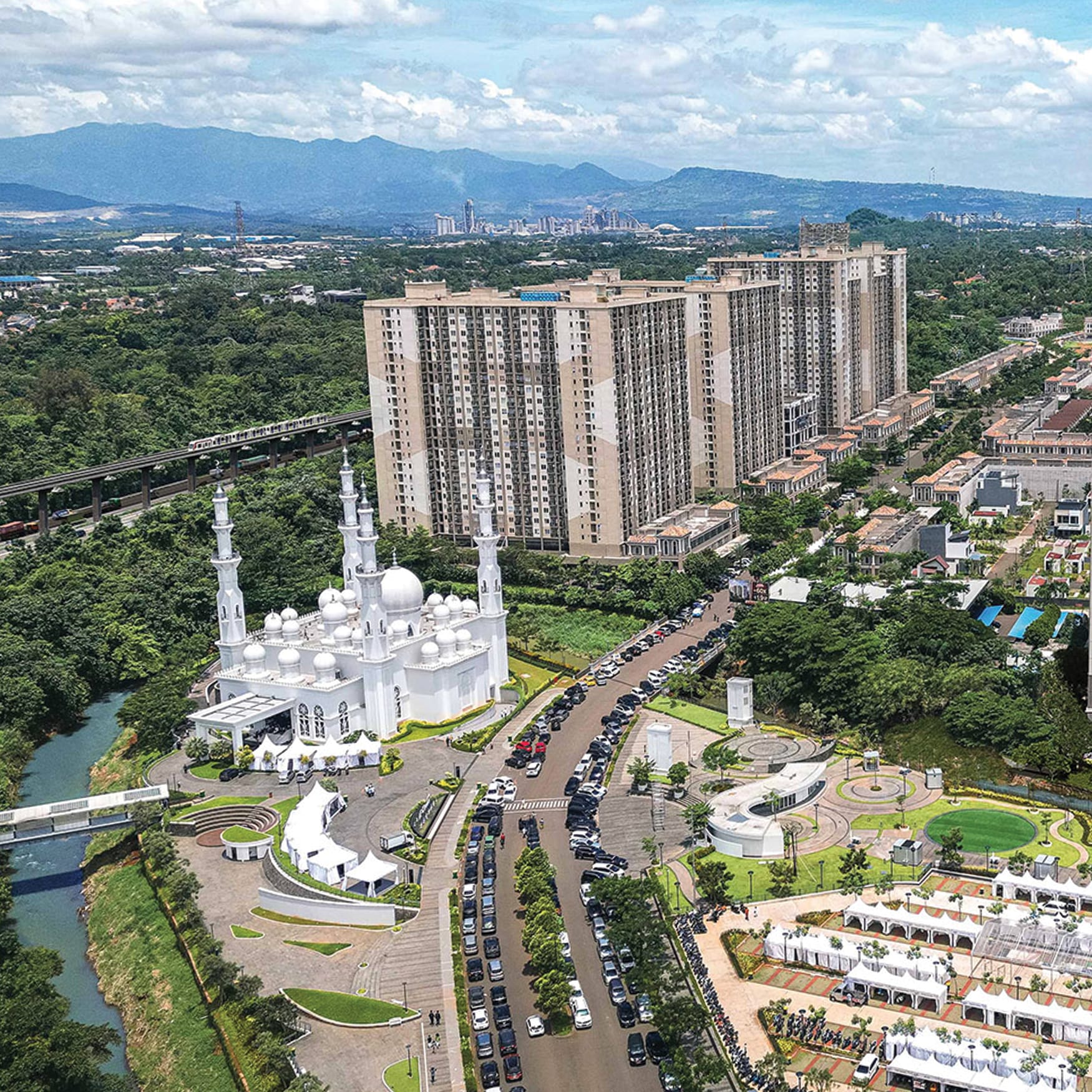 Aerial view of a landscaped urban district in Indonesia featuring a white mosque, residential towers, tree-lined roads, public plazas, and a tropical cityscape surrounded by mountains.