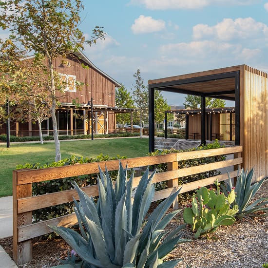 Rustic barn-style building beside a modern black steel and wood pavilion with string lights, separated by a horizontal wood slat fence, with agave and prickly pear cactus landscaping in the foreground and a lawn between the structures.