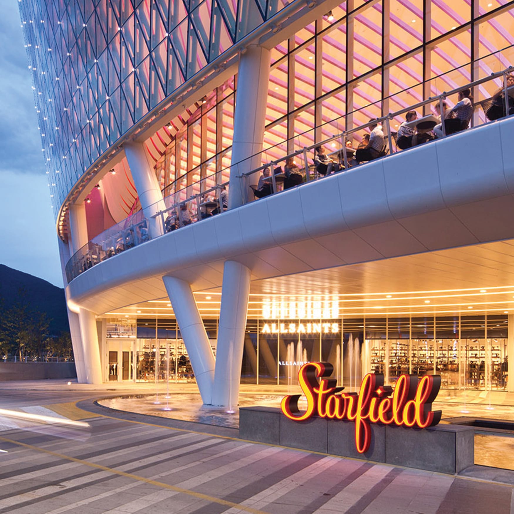 Illuminated entrance of Starfield Hanam shopping mall in South Korea at night, featuring a glowing red logo, modern glass architectural facade, and outdoor seating balcony.