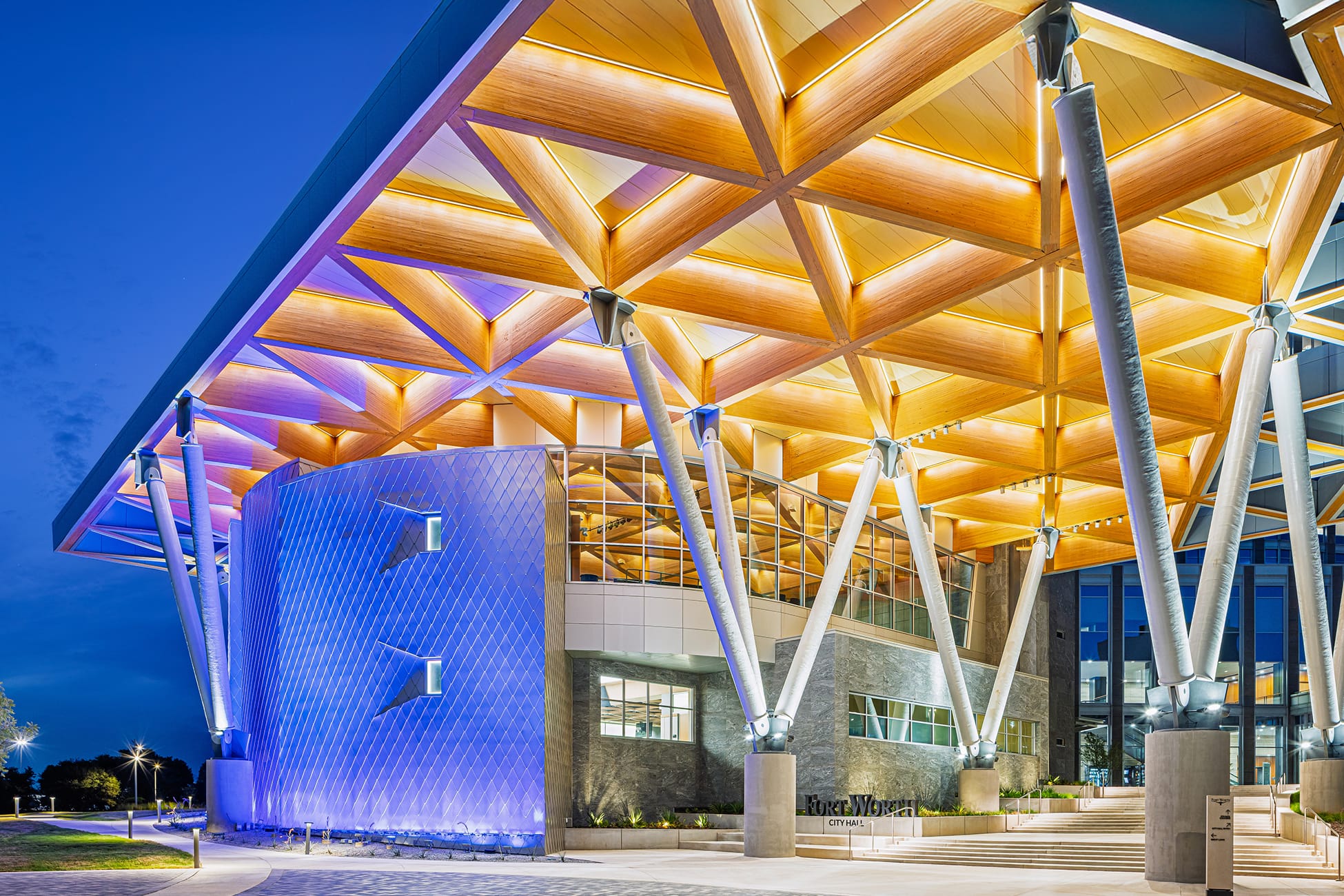 Fort Worth City Hall dramatic dusk exterior showcasing illuminated wood lattice canopy, branching white structural columns, blue-lit diamond-pattern Council Chamber facade, and Fort Worth City Hall monument sign