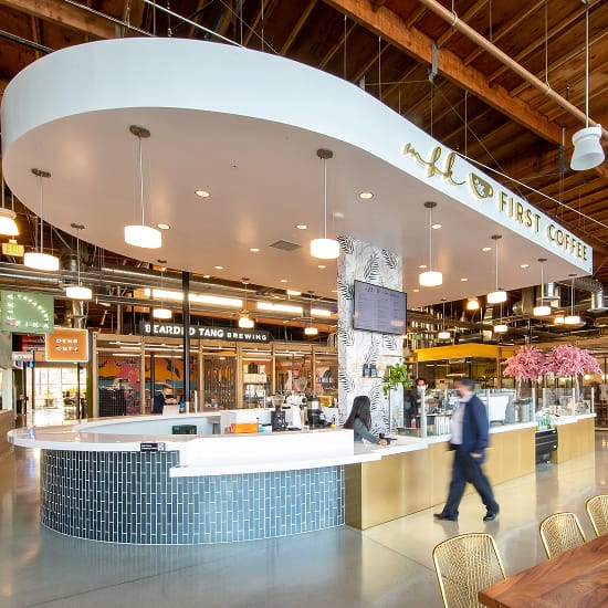 Interior of the "First Coffee" bar inside Rodeo 39 Public Market, featuring a curved white oval soffit with gold lettering, a tiled counter with brass base, tropical leaf wallcovering, and pendant lighting beneath an exposed wood beam ceiling, with Bearded Tang Brewing and other vendor stalls visible in the background.