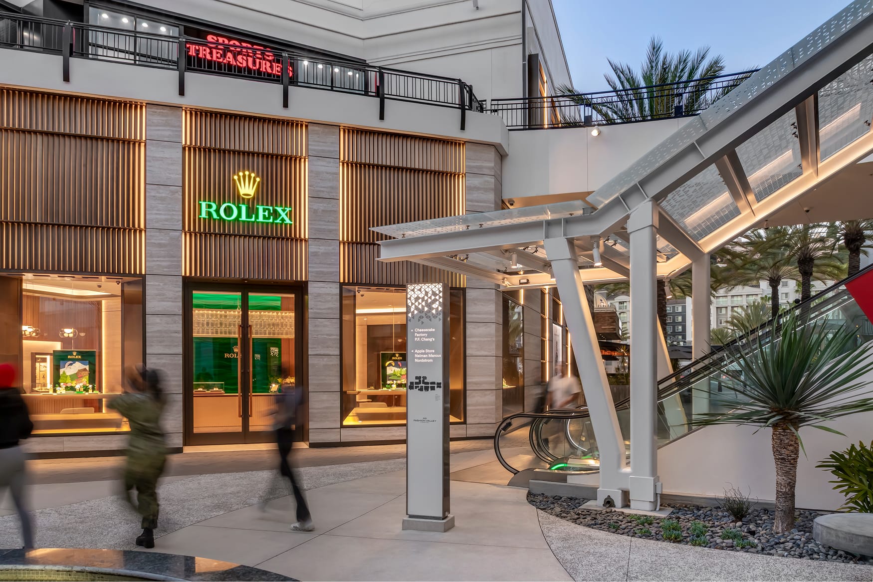Exterior view of the Rolex boutique at Fashion Valley Mall, featuring warm wood-toned slat facade and illuminated green signage, with a wayfinding pylon, modern white canopy structure, escalator, and palm trees in the foreground at dusk.