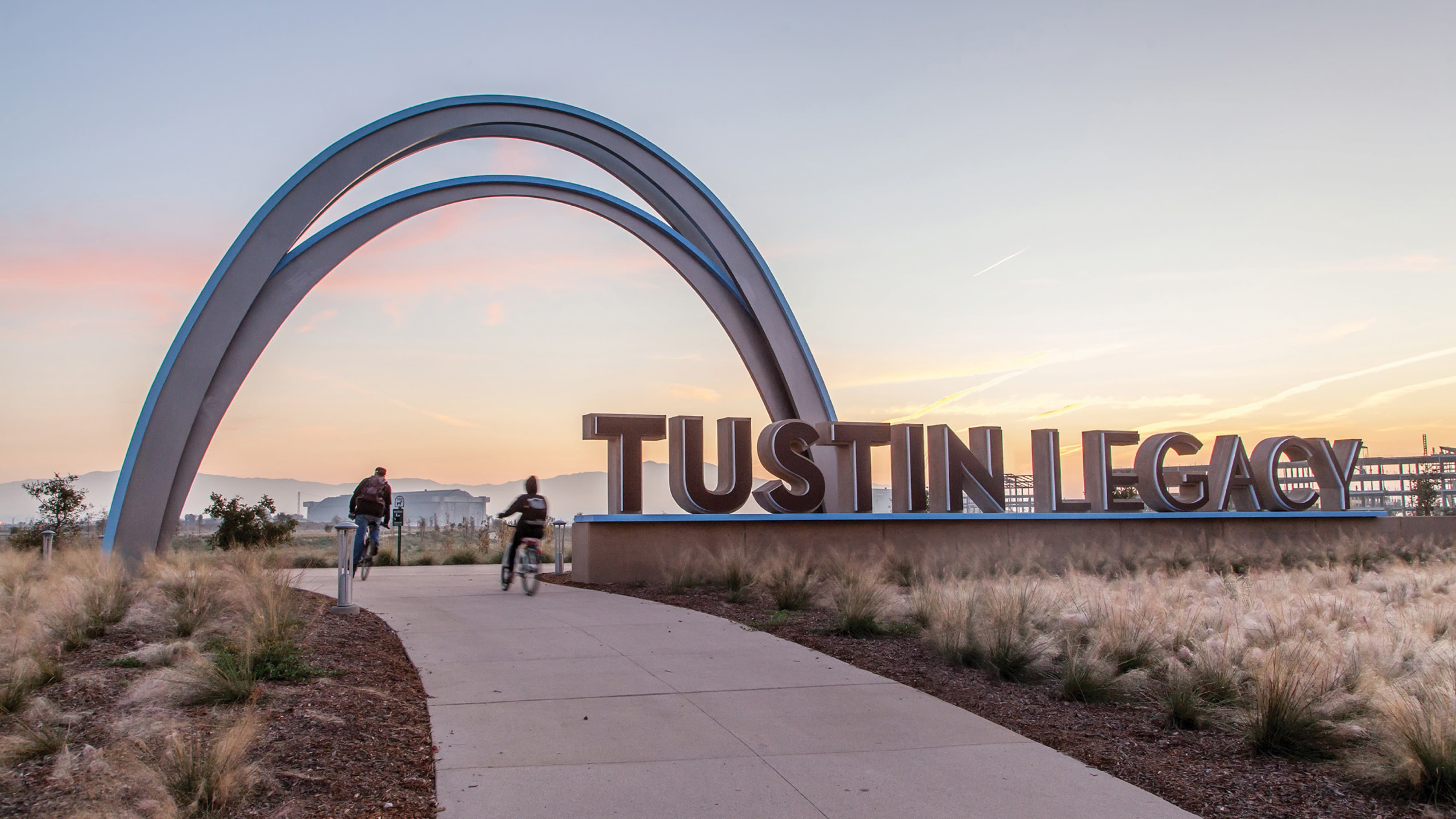Monumental 'Tustin Legacy' sign with dimensional letters beneath a curved trio of archways, set along a paved multi-use path lined with native grasses; two cyclists ride toward the sign at sunset, with distant hills and cranes visible in the background.