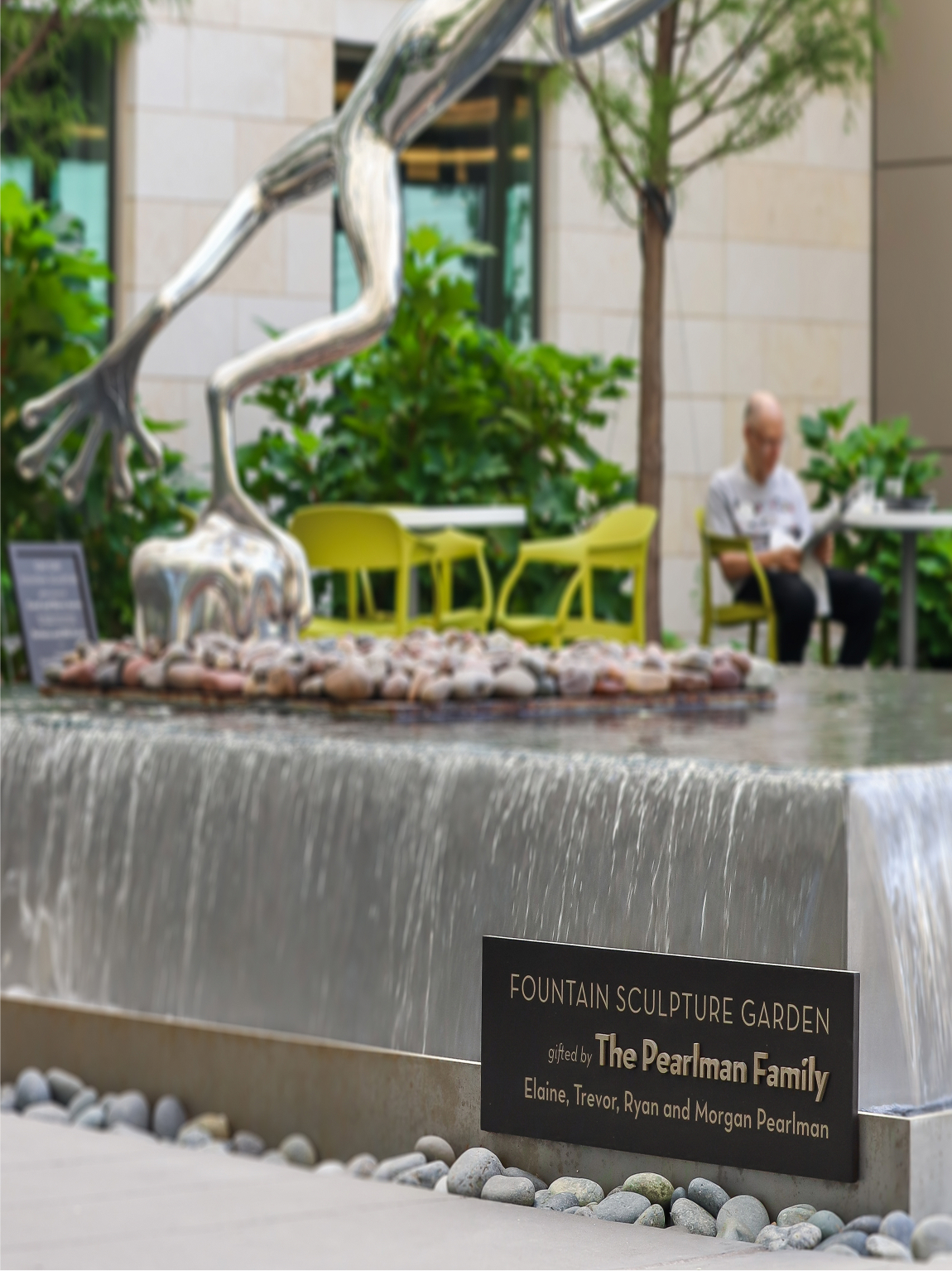 Fountain Sculpture Garden with a chrome running figure sculpture atop a cascading water wall, a plaque reading 'gifted by The Pearlman Family, Elaine, Trevor, Ryan and Morgan Pearlman,' and a courtyard seating area with yellow chairs in the background.
