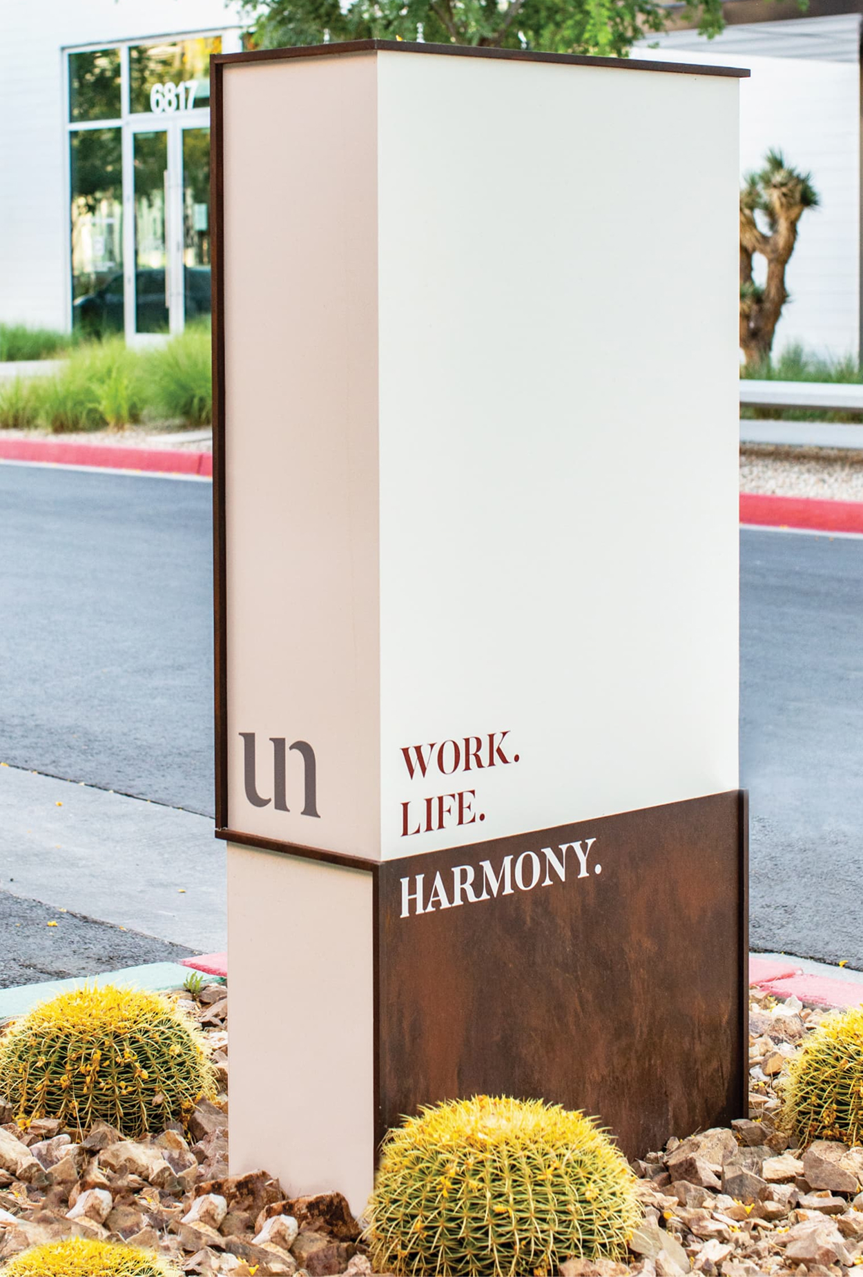 A tall freestanding monument sign for Uncommons at a street corner in Las Vegas. The sign features a cream upper panel with the "un" logo mark and the words "Work. Life." in dark red, transitioning to a Corten steel base with "Harmony." in white. Golden barrel cacti and rock landscaping surround the base, with a commercial building at 6817 and desert plantings visible in the background.