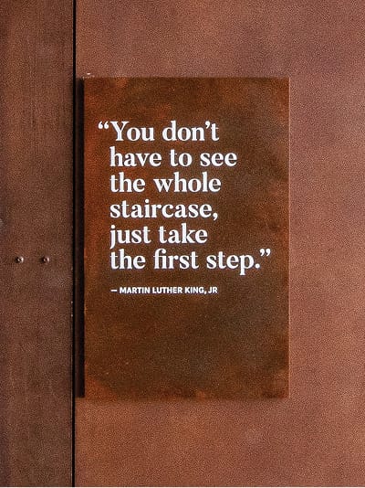 A dark plaque mounted on a Corten steel wall displays a quote in white serif type: "You don't have to see the whole staircase, just take the first step." attributed to Martin Luther King, Jr.