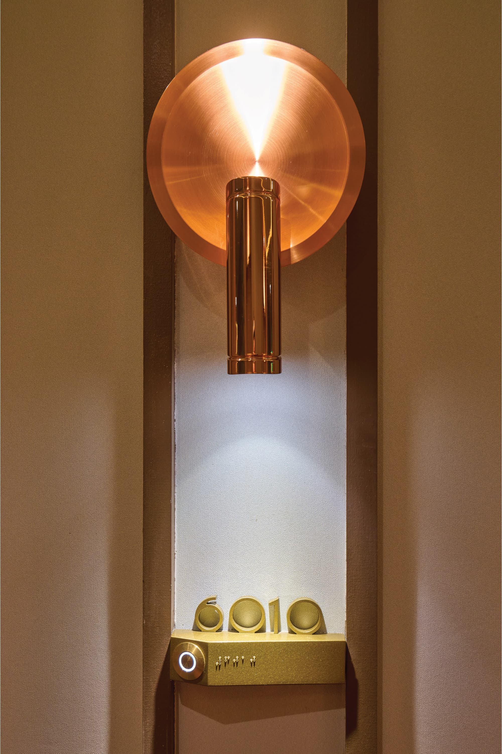 Close-up detail of a brushed brass ADA-compliant room number sign with Braille and tactile characters beneath a warm copper wall sconce light fixture in a hotel corridor at Pier Sixty-Six luxury waterfront resort in Fort Lauderdale, Florida — RSM Design wayfinding and signage design, master sign programs, specialty graphics, and branded environments for hospitality and waterfront destinations.