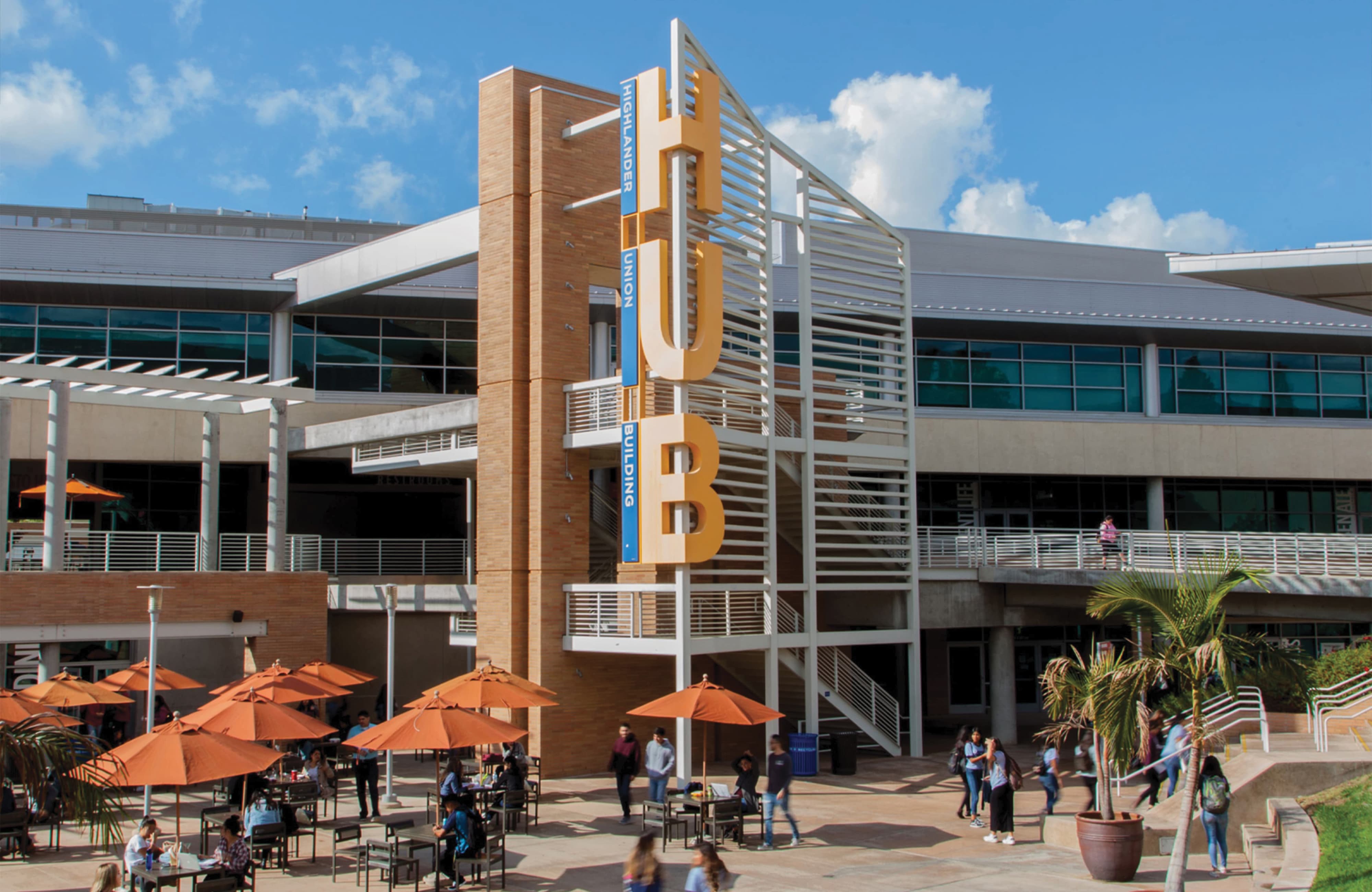 Large orange 'HUB' letter sculpture with 'Highlander Union Building' signage mounted on a brick tower, in front of a busy campus courtyard with orange umbrella-shaded seating, palm trees, and students walking between buildings.