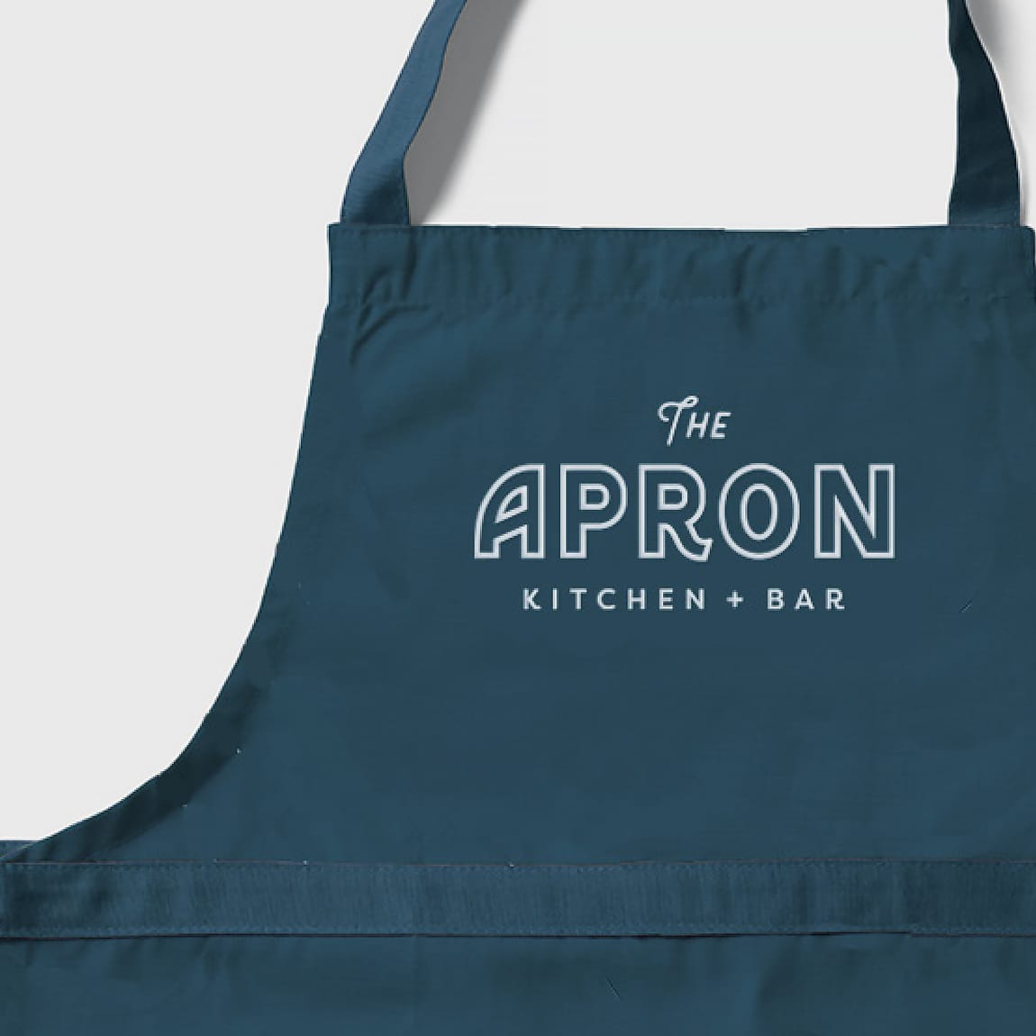 The Apron Kitchen + Bar branded staff apron in teal with white logo featuring script and bold outlined typography, restaurant uniform design at Omni PGA Frisco