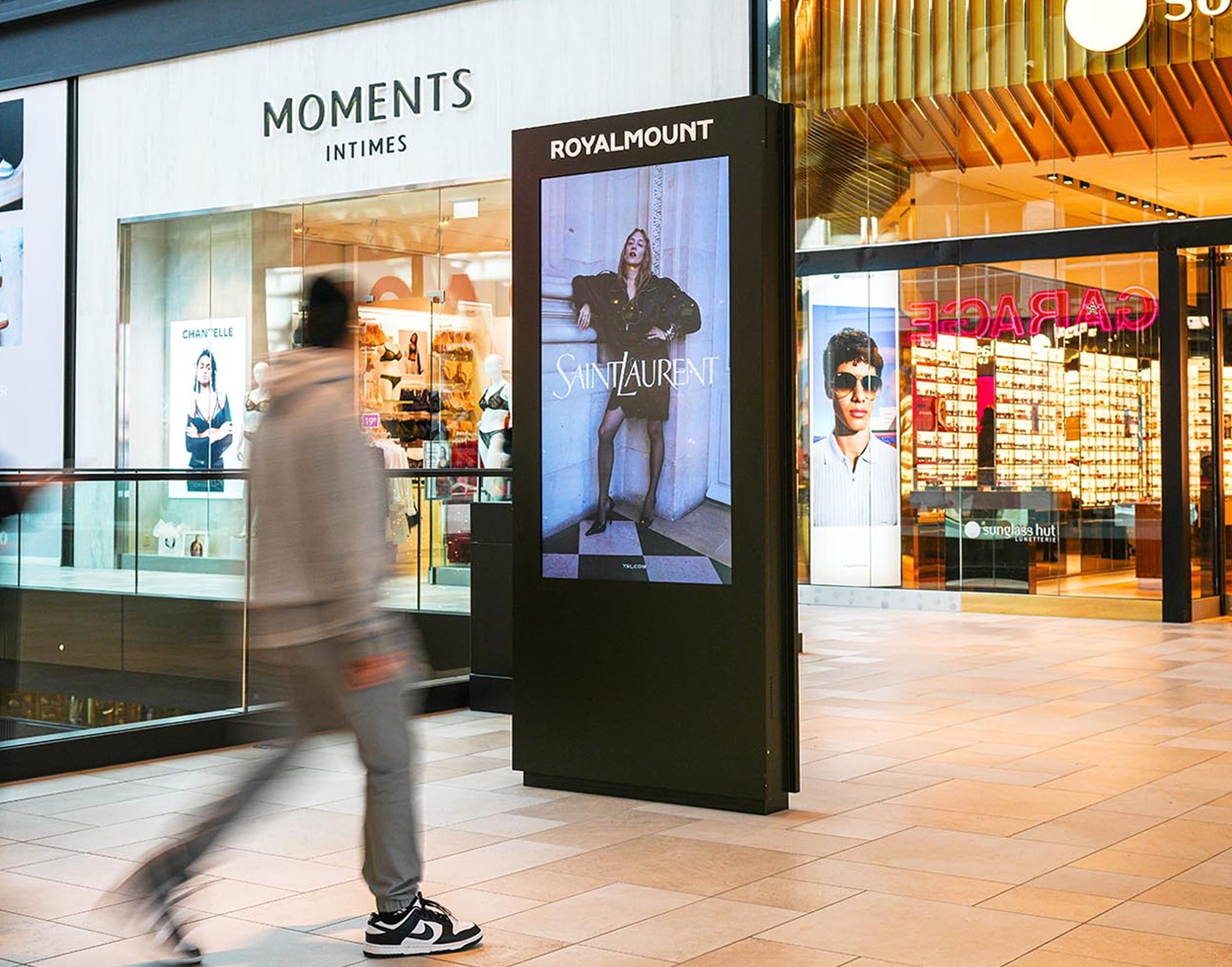 Digital Royalmount-branded advertising column displaying a Saint Laurent campaign, blurred shopper walking past storefronts in the concourse — digital signage design by RSM Design