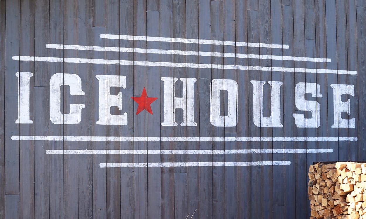 Close-up of hand-painted Ice House mural on weathered wood plank wall featuring bold white stencil lettering, red star logo, and stacked firewood at PGA Frisco
