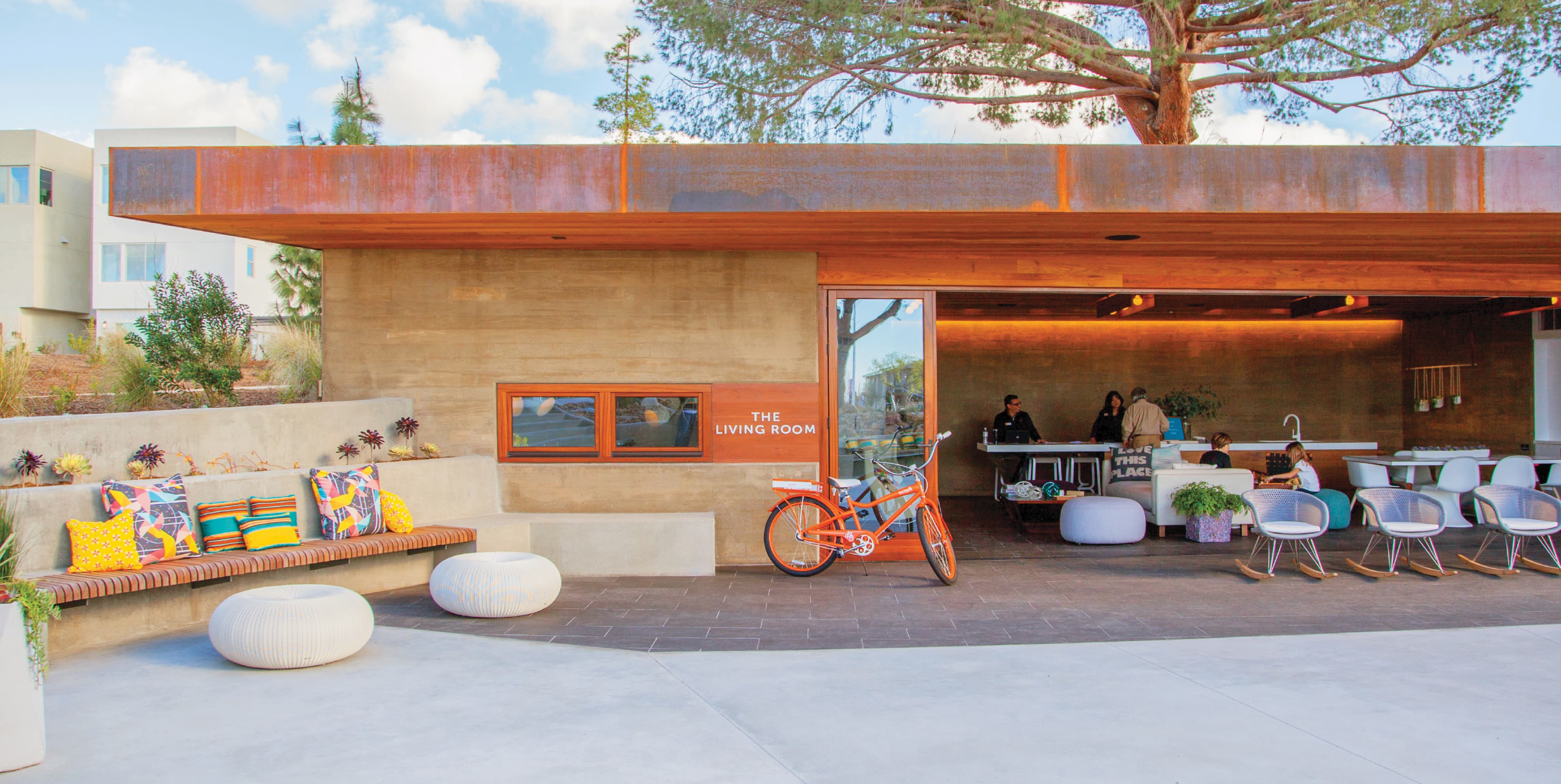 RSM Design building identity signage and placemaking for The Living Room community amenity at FivePoint Great Park Neighborhoods in Irvine, California, featuring dimensional white entry signage integrated into a warm wood and rammed earth community pavilion facade, with an orange branded cruiser bicycle, colorful outdoor seating, patterned pillows, and residents gathered at a community counter in the background, illustrating how environmental graphic design, visual identity, experiential graphics, and placemaking work together to create an inviting, human-centered branded environment that transforms a community amenity space into a genuine neighborhood gathering place for residential community and park markets.