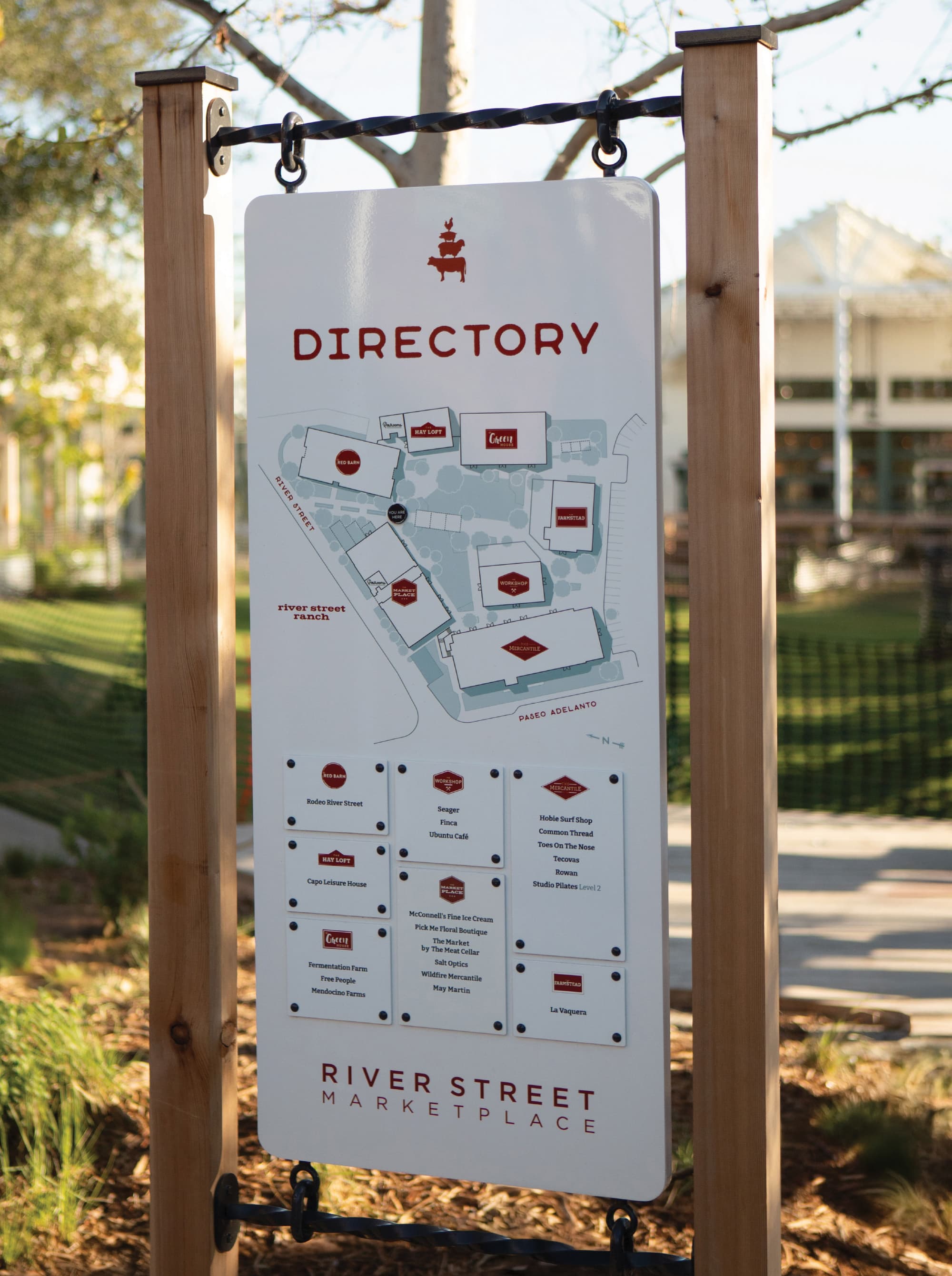River Street Marketplace directory sign suspended from a rustic wood and wrought iron post frame, displaying a site map and tenant listings including McConnell's Fine Ice Cream, Mendocino Farms, Hobie Surf Shop, and Seager, showcasing wayfinding and signage and environmental graphic design for a mixed-use waterfront retail environment by RSM Design.