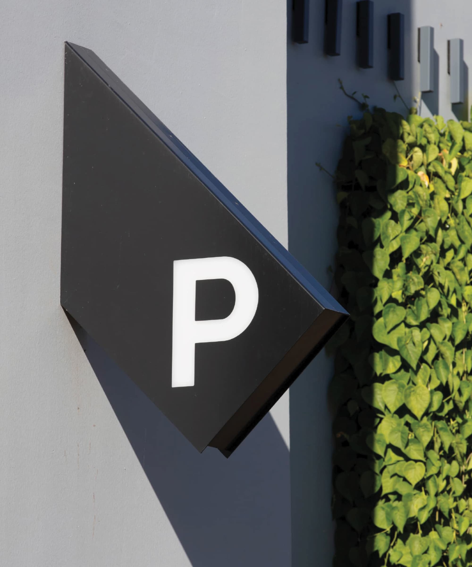 Angular black diamond-shaped 'P' parking sign mounted on a gray building facade, beside a green living wall of climbing vines.
