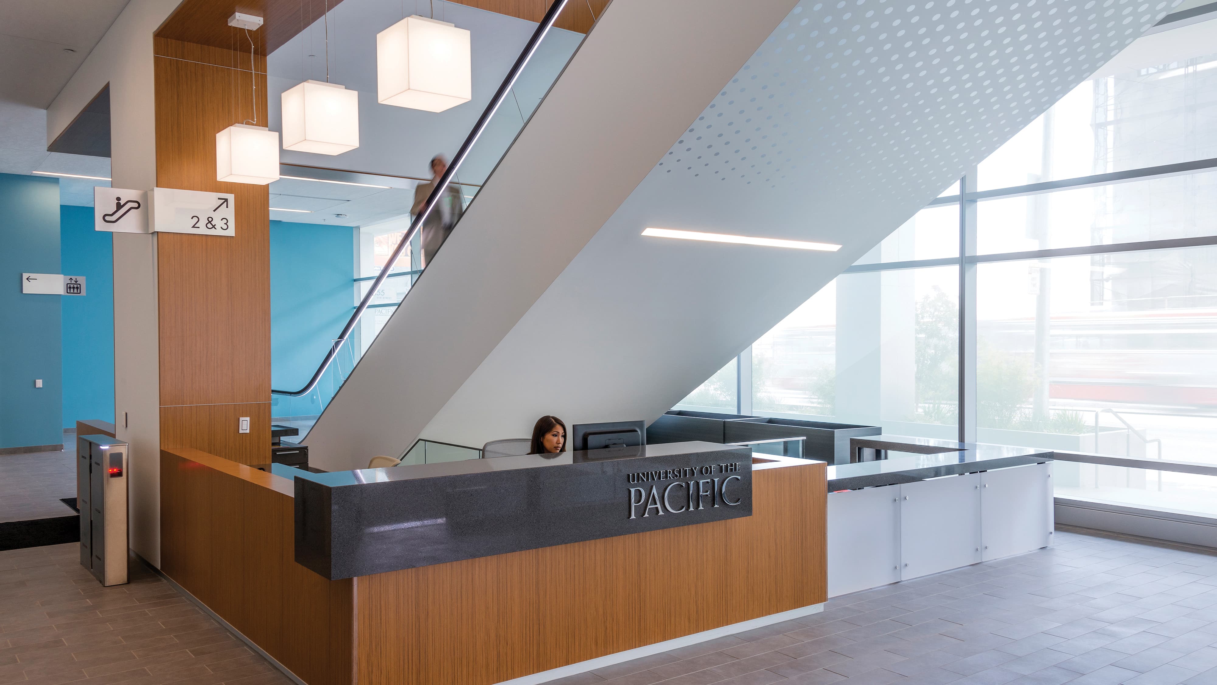 University of the Pacific San Francisco Campus interior lobby reception desk with dimensional institutional logotype on a dark granite face panel, escalator level identification signage directing to floors 2 and 3, and elevator directional sign on a bold blue accent wall — RSM Design branding and visual identity, wayfinding and signage design, master sign programs, and environmental graphic design for education and health and wellness destinations.