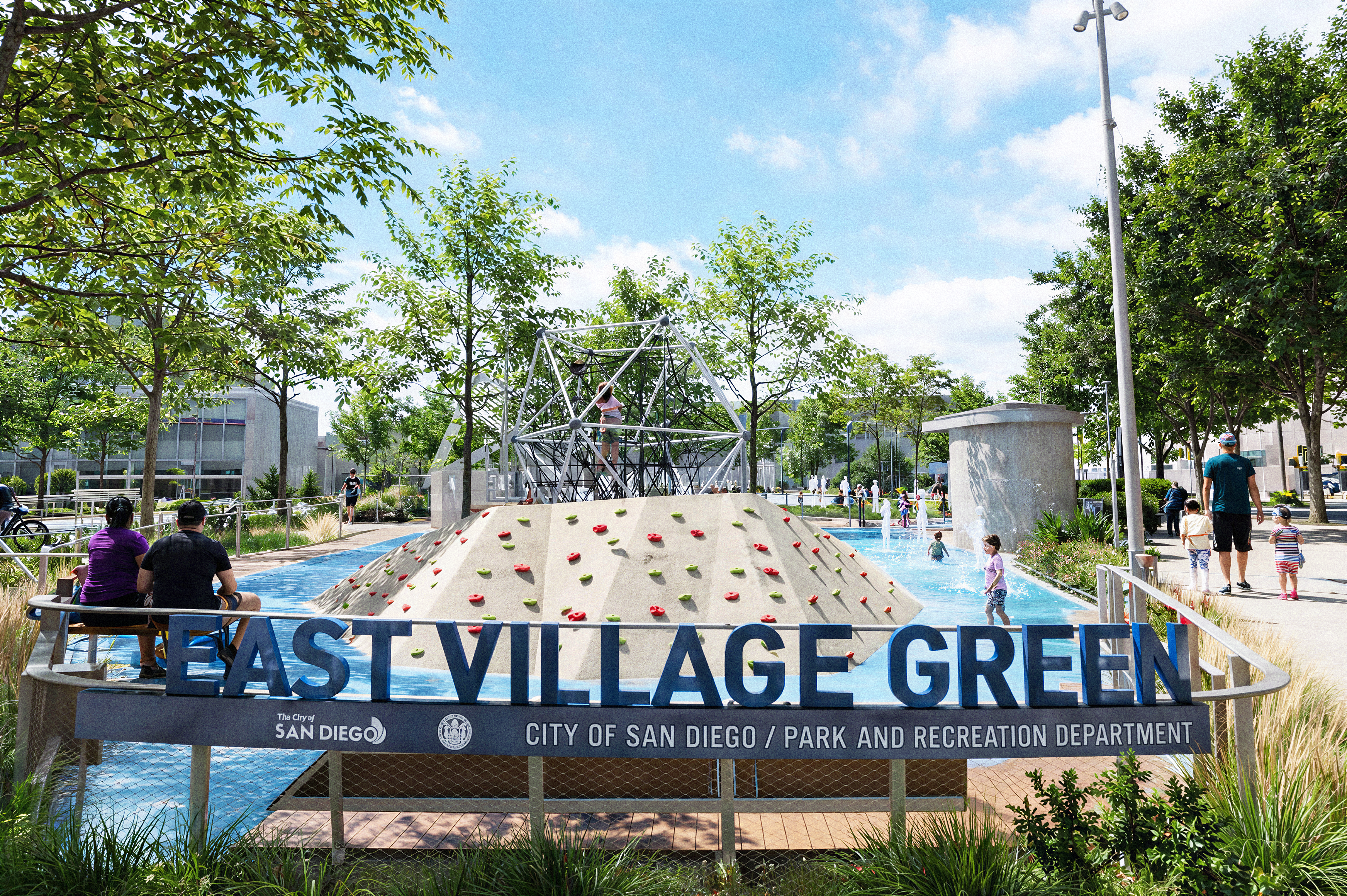 Large 'East Village Green, City of San Diego / Park and Recreation Department' sign on a railing overlooking a climbing dome and rock wall pyramid, with children playing in a splash pad and families walking through the tree-lined park.