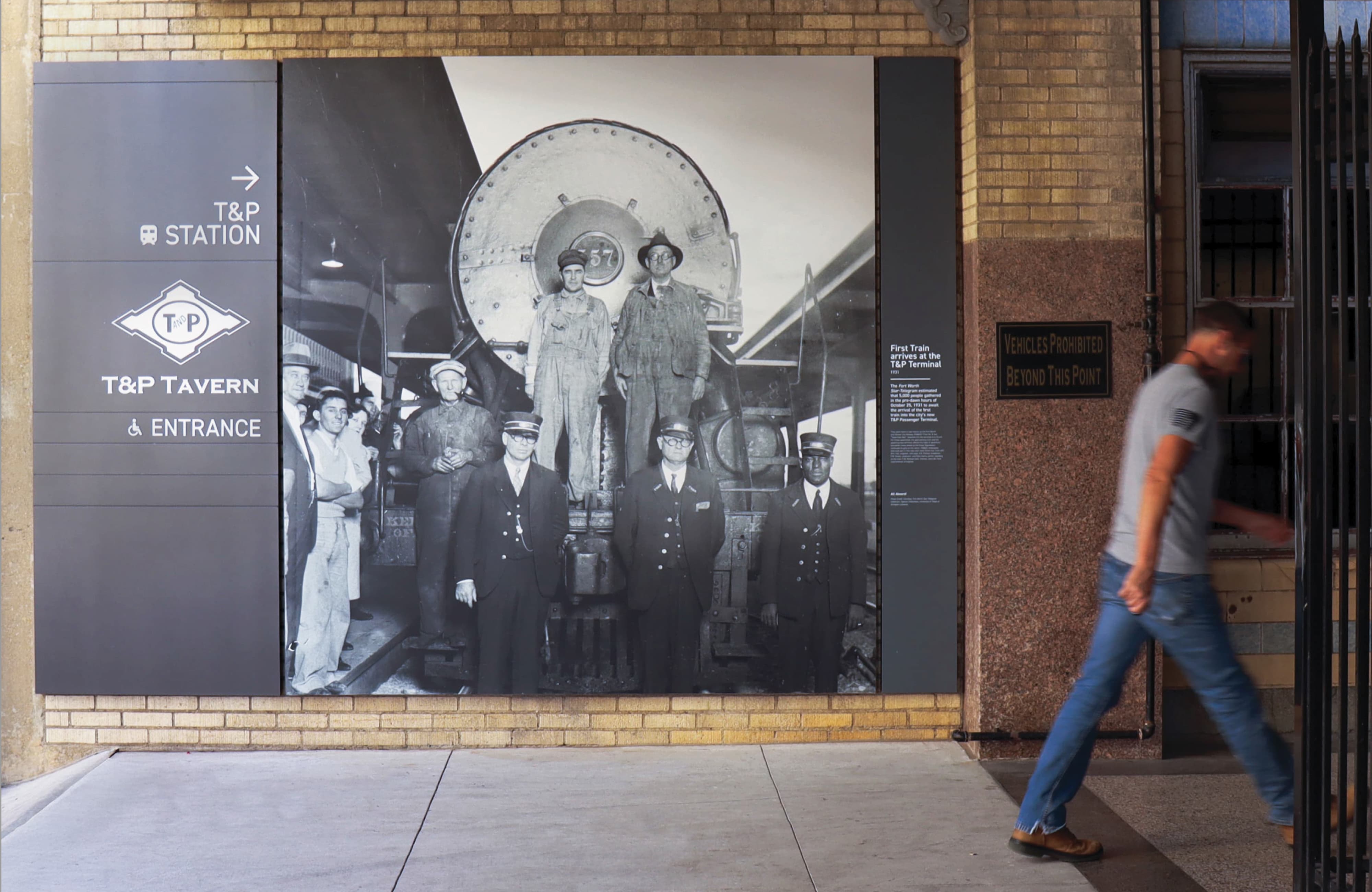 Exterior wall at T&P Station featuring a large-scale historic black-and-white photographic mural of the first train's arrival alongside wayfinding signage directing visitors to T&P Station and T&P Tavern entrance, blending experiential graphics, environmental graphic design, and placemaking for a mixed-use transit development by RSM Design