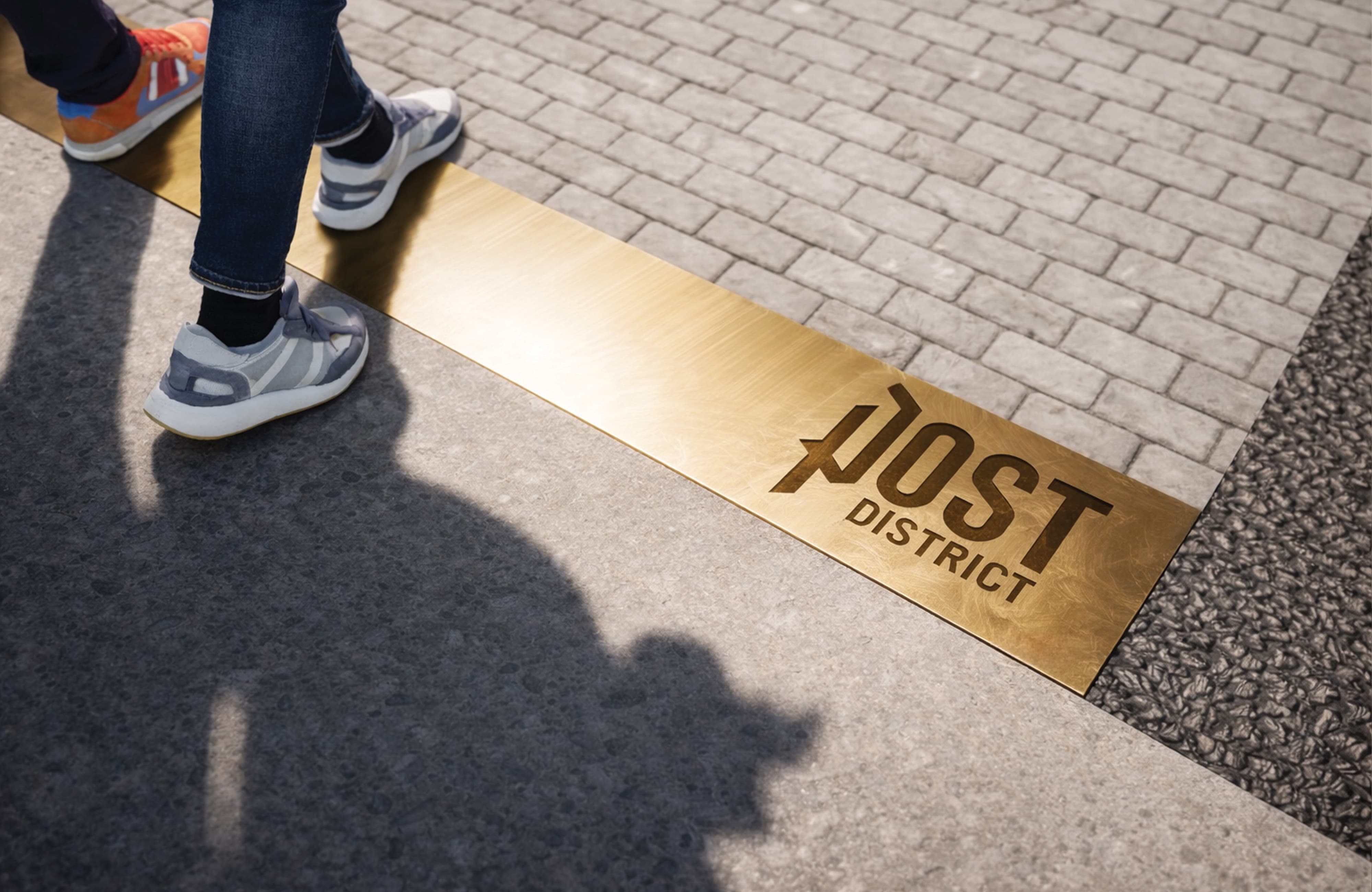 Pedestrian-level view of a brass inlaid ground graphic featuring the Post District logo embedded in a paver streetscape — a specialty graphic design and placemaking detail by RSM Design for a mixed-use residential community.