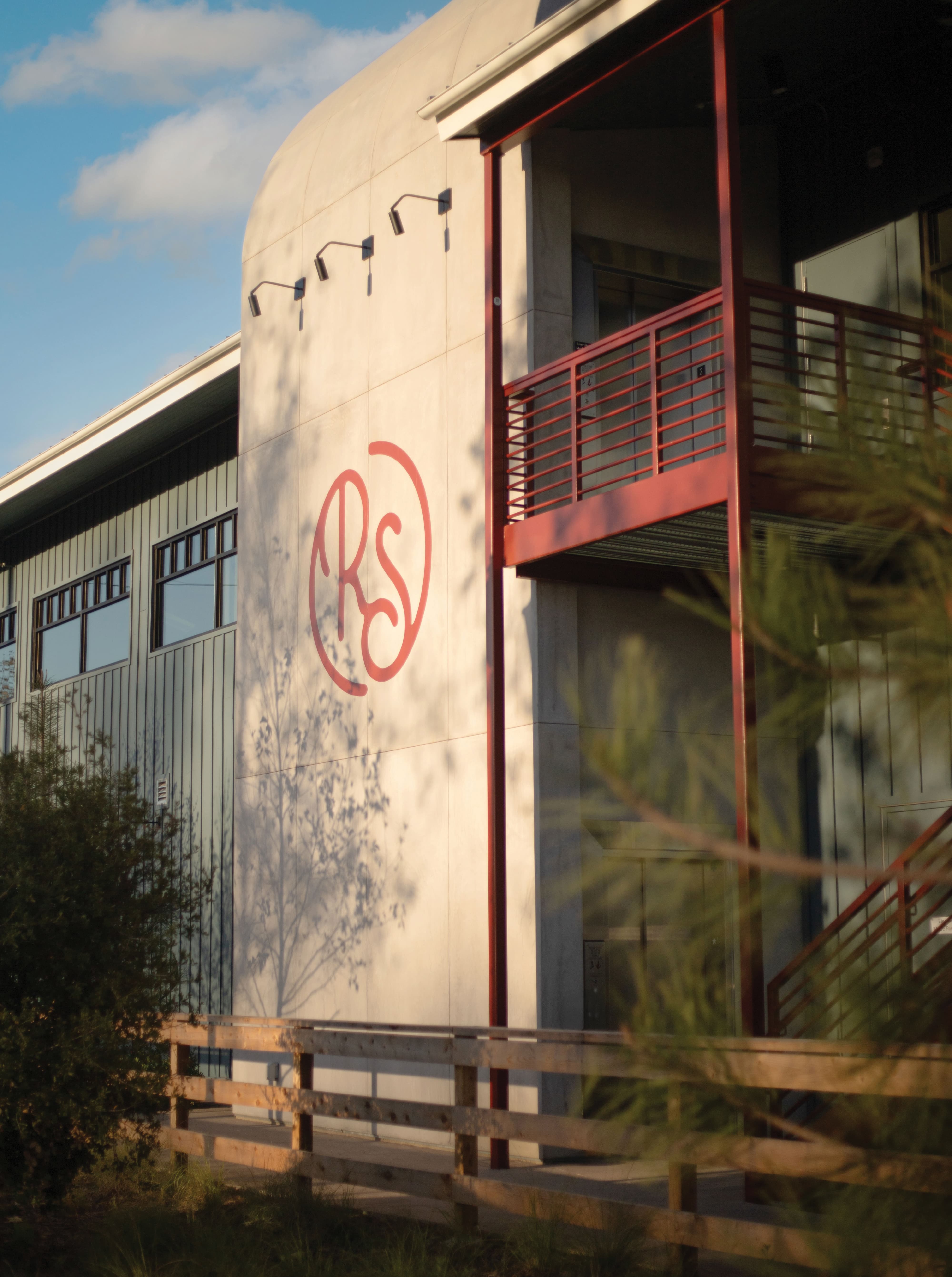 RSM Design large-scale exterior logo application for River Street Marketplace, featuring the RS monogram rendered in hand-drawn script directly on a curved architectural wall surface at golden hour, integrating visual identity and experiential graphics seamlessly into the building architecture to establish a distinctive landmark and sense of place for a mixed-use retail and streetscape environment.