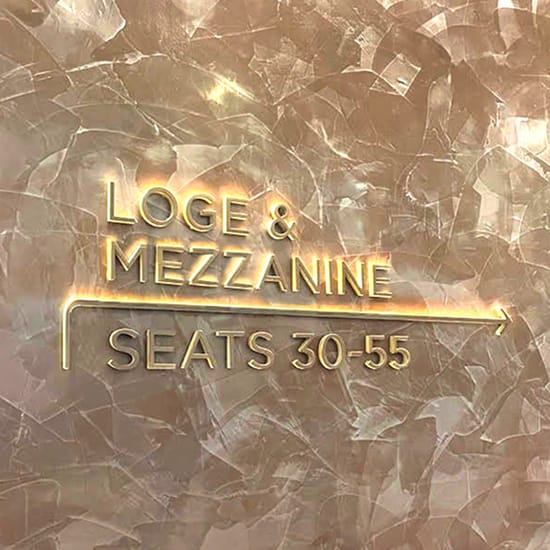 Illuminated gold dimensional lettering wayfinding sign reading "Loge & Mezzanine Seats 30-55" with directional arrow mounted on a textured rose gold wall at Buddy Holly Hall of Performing Arts and Sciences, Lubbock Texas