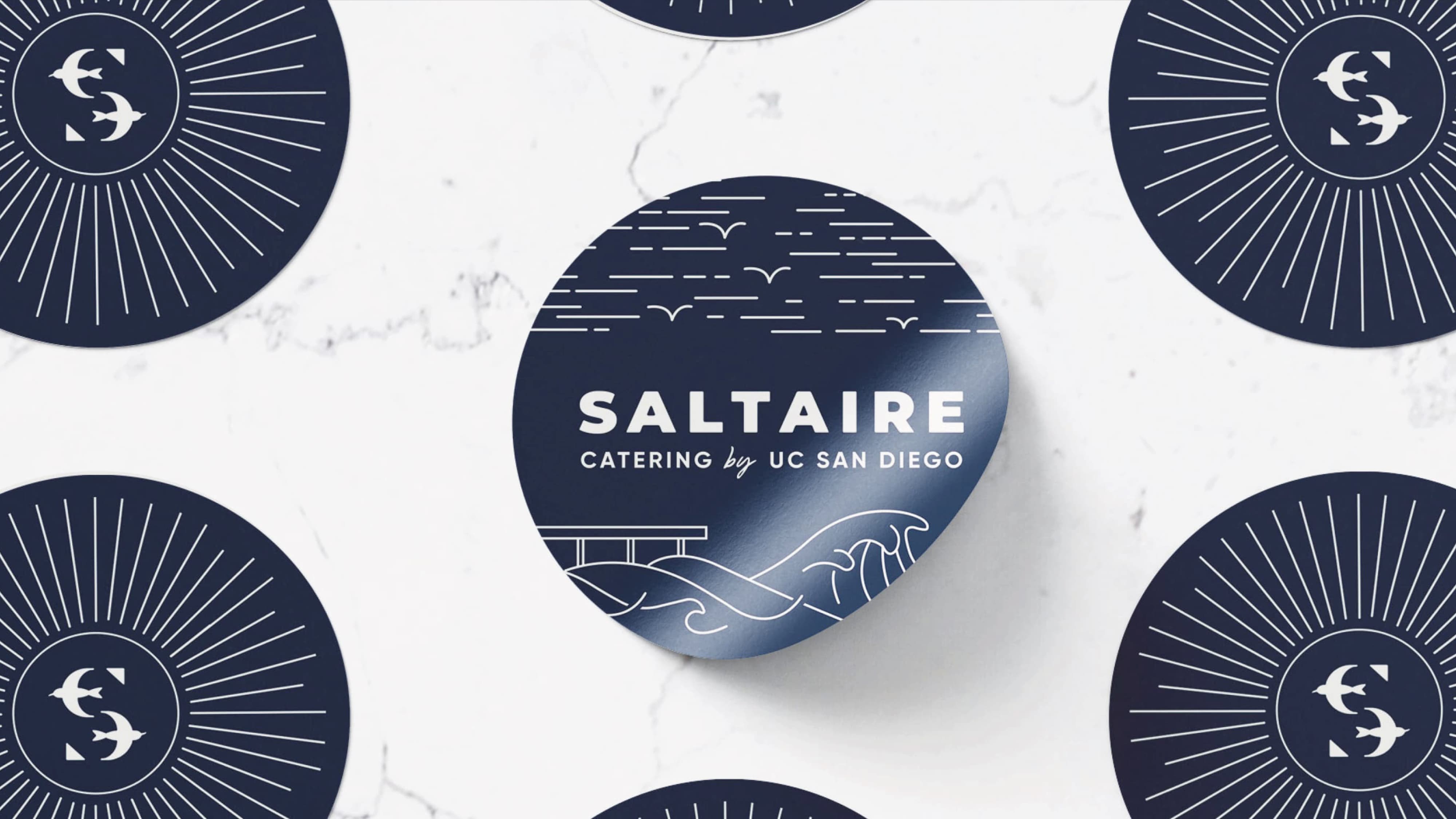 Saltaire Catering by UC San Diego branded coasters in navy featuring a coastal illustration with waves, terns, and a pier alongside the sunburst lettermark pattern on a marble surface, showcasing specialty graphics and print collateral design for a food and beverage and education market client by RSM Design.
