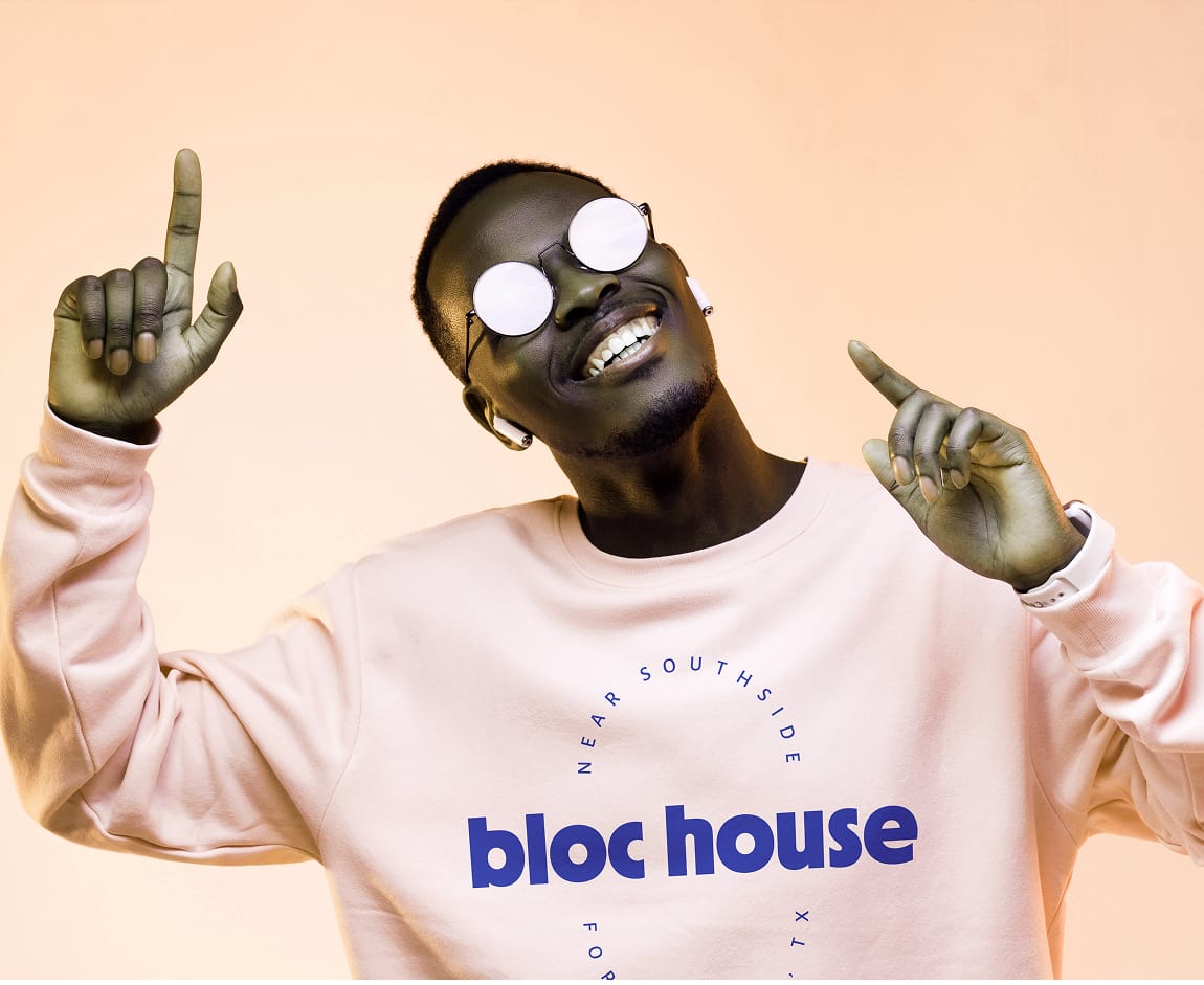A smiling man wearing round mirrored sunglasses and a blush pink crewneck sweatshirt printed with the "bloc house" wordmark in bold blue type and the text "Near Southside, Fort Worth, TX" arched above, pointing upward with both hands against a warm peach background.