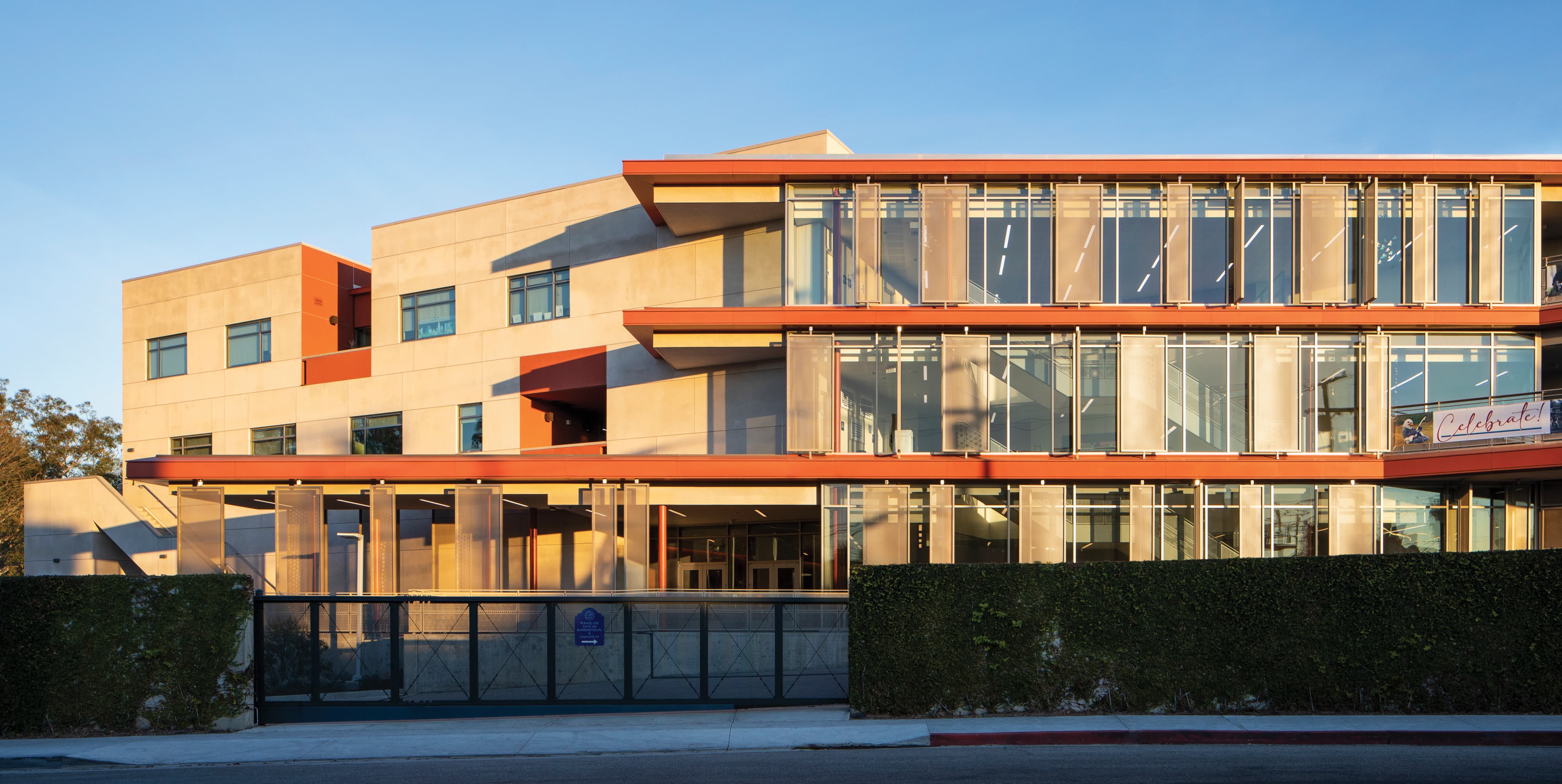 A wide exterior view of a multi-story educational campus building bathed in warm golden hour light, featuring red architectural banding, expansive curtain wall glazing, a gated pedestrian entry with a blue wayfinding sign panel, and a "Celebrate!" banner visible through the upper floor windows — illustrating RSM Design's environmental graphic design and wayfinding and signage design services integrated cohesively into the architecture and placemaking of the education market