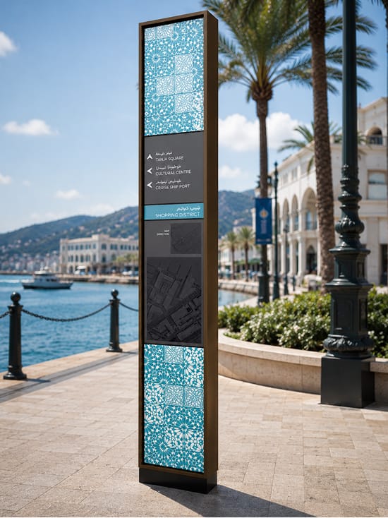 Rendering of a tall wayfinding pylon with teal geometric tile patterns and bilingual directional listings for Tanja Square, Cultural Centre, Cruise Ship Port, and Shopping District, standing on a waterfront promenade beside palm trees and Mediterranean-style buildings.