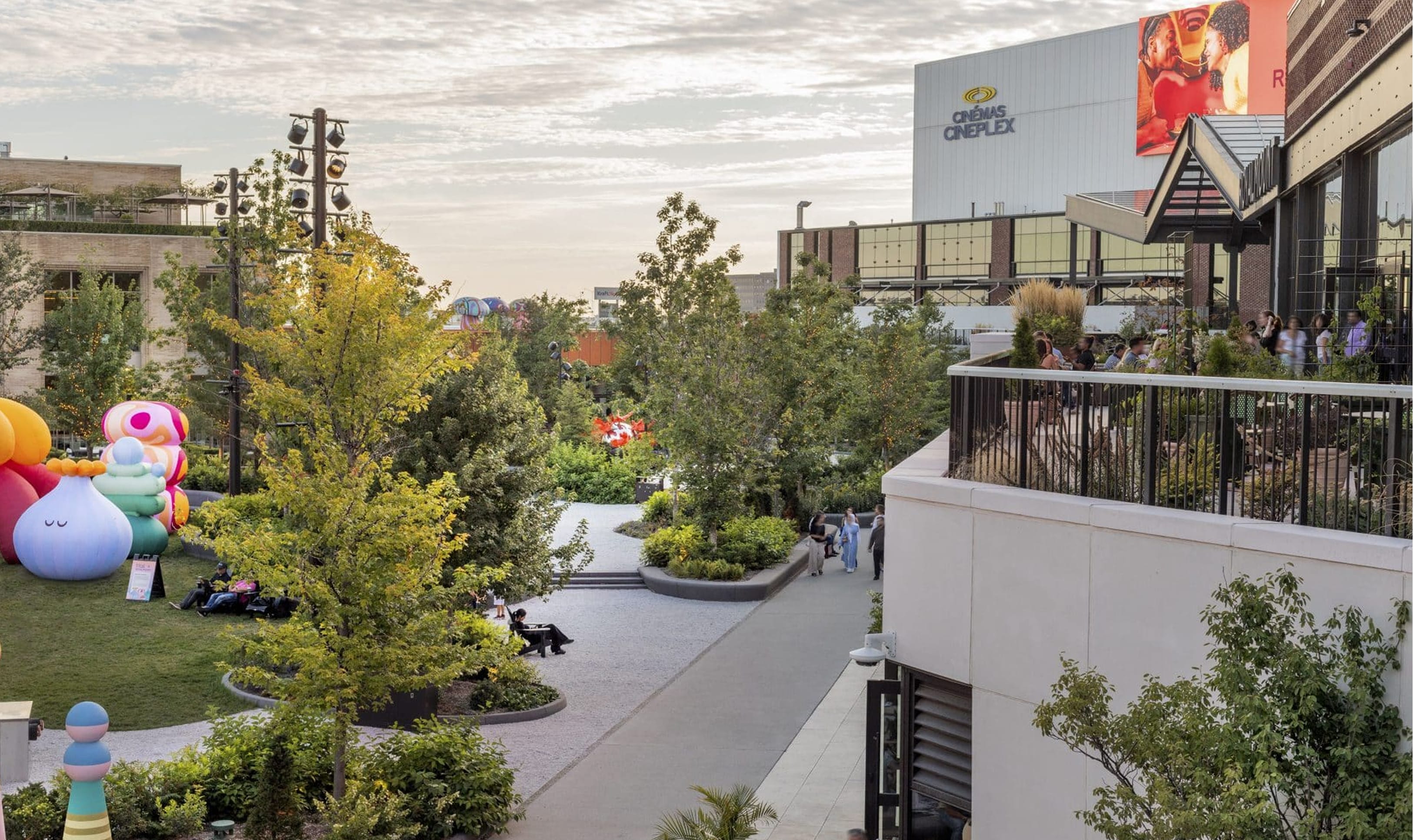 Overhead view of a landscaped plaza at Royalmount with whimsical inflatable sculptures on the lawn, a rooftop terrace with diners, and the Cinémas Cineplex building in the background at dusk — placemaking design by RSM Design