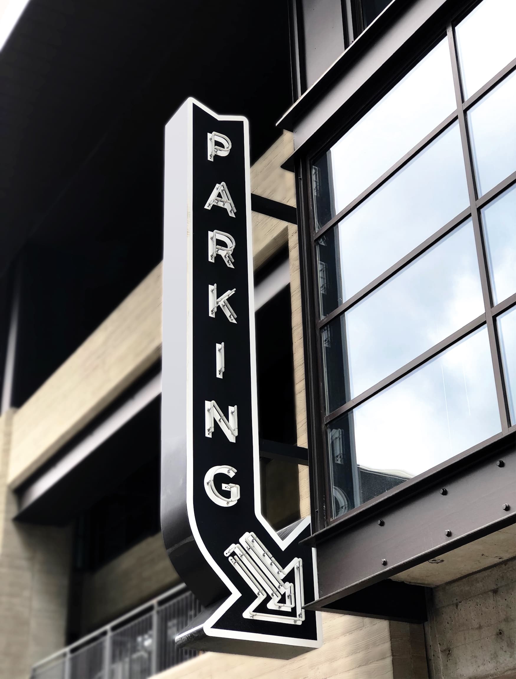 Black vertical letters "Parking" blade sign in the shape of an arrow with a curved arrow head and white graphics and outlining with neon tubing for illumination. Signage and wayfinding for Fifth + Broadway in Nashville, Tennessee by RSM Design