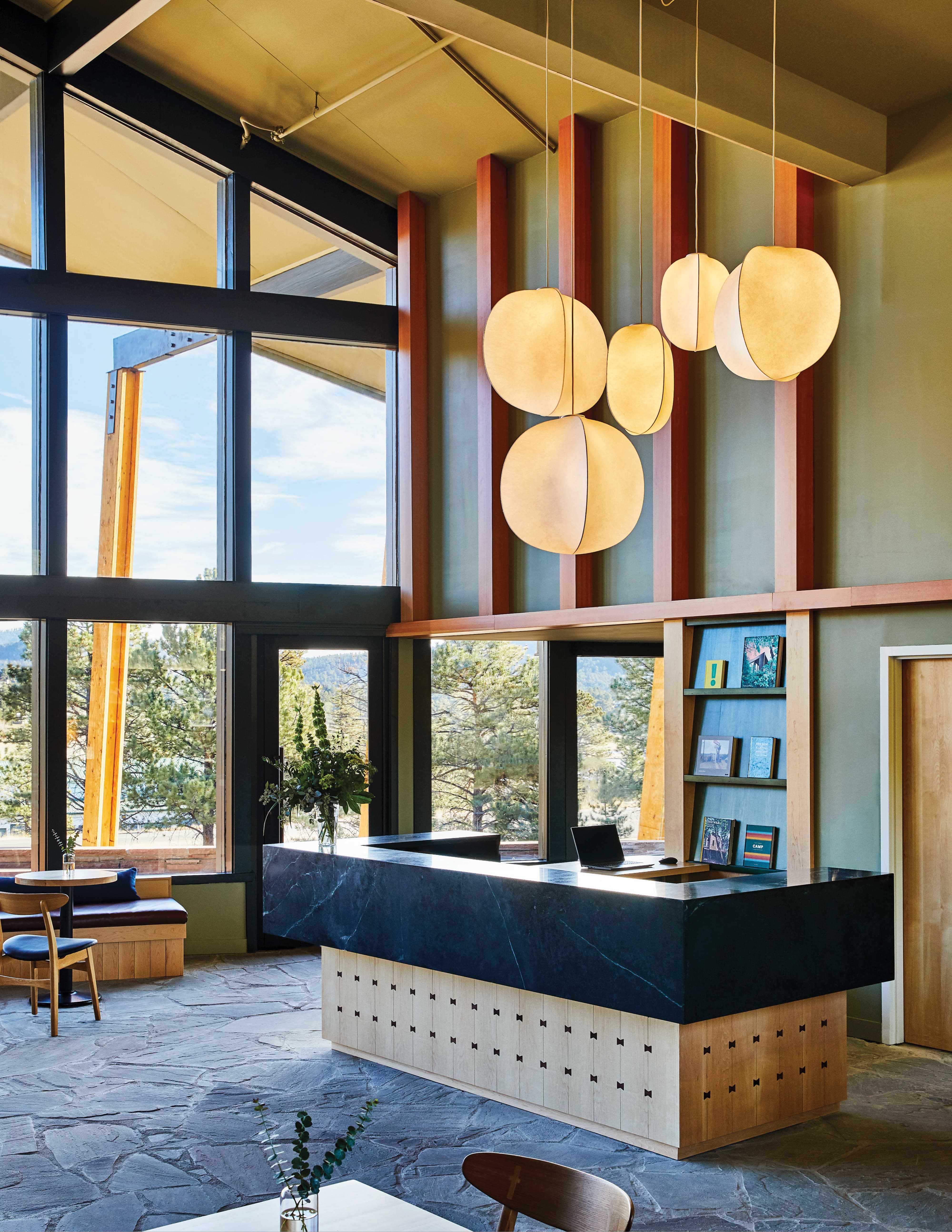 Front desk lobby interior at Trailborn Rocky Mountains Outpost featuring a dark marble reception counter, sculptural pendant lighting, colorful vertical striped wall treatment, and floor-to-ceiling windows overlooking pine trees, showcasing placemaking and experiential design for a hospitality environment by RSM Design.
