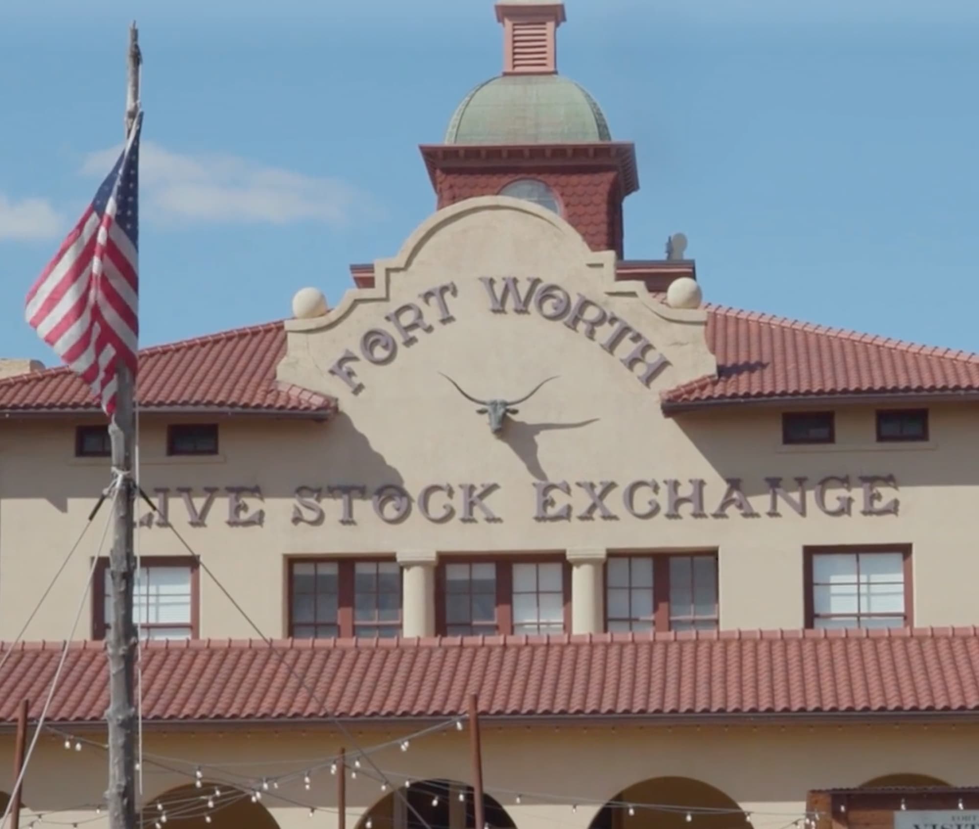 A building in historical Spanish colonial style architecture, with beige stucco walls and red clay tile roofs. Applied lettering on the façade reads “Fort Worth / Live Stock Exchange” in all capital letters in a swashed, Tuscan serif font. A cast metal bull’s head sits between the two lines of text. In front of the building, an American flag hangs from a natural wood pole.