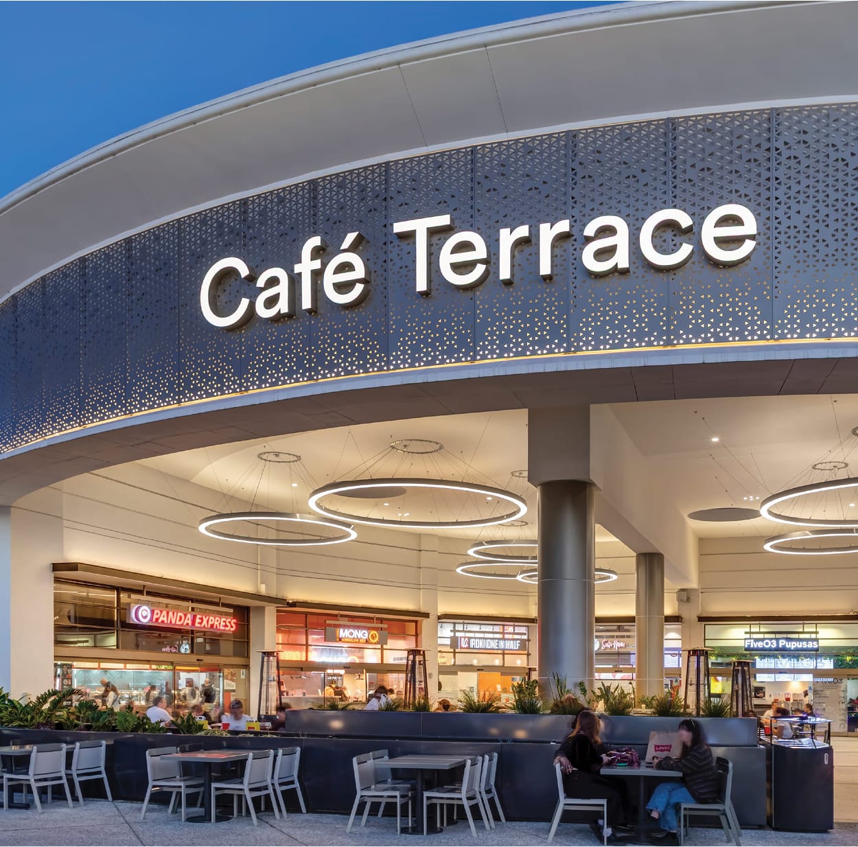 Exterior view of the Café Terrace food court at Fashion Valley Mall at dusk, featuring a curved perforated metal façade with backlit signage, circular pendant lighting, and outdoor seating with diners, with Panda Express, Mongo, and FiveO3 Pupusas visible inside.