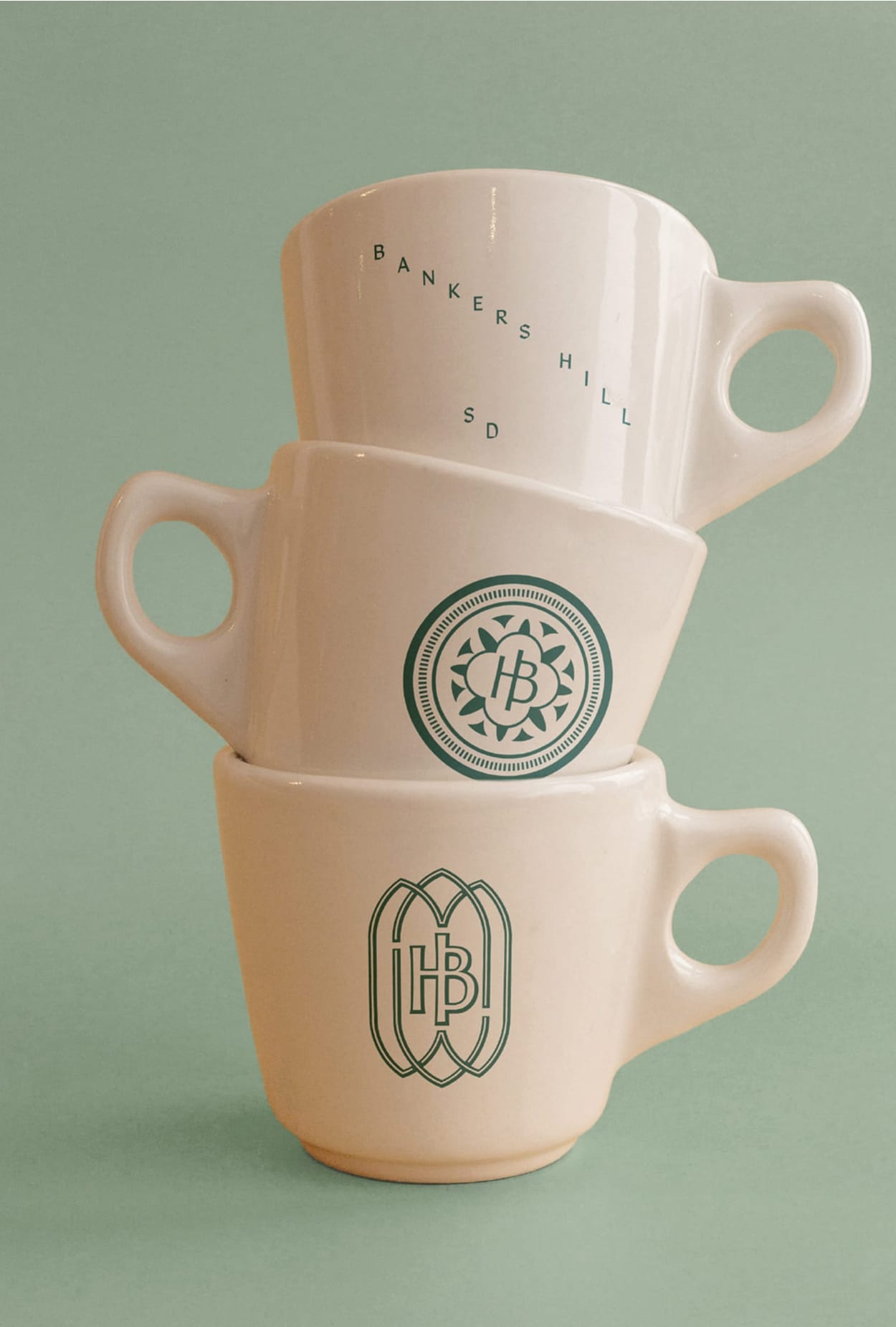 Stacked ceramic coffee mugs featuring Heavenly Bodega branding in San Diego, including HB monogram logos and “Bankers Hill SD” typography in deep green on a soft neutral background.