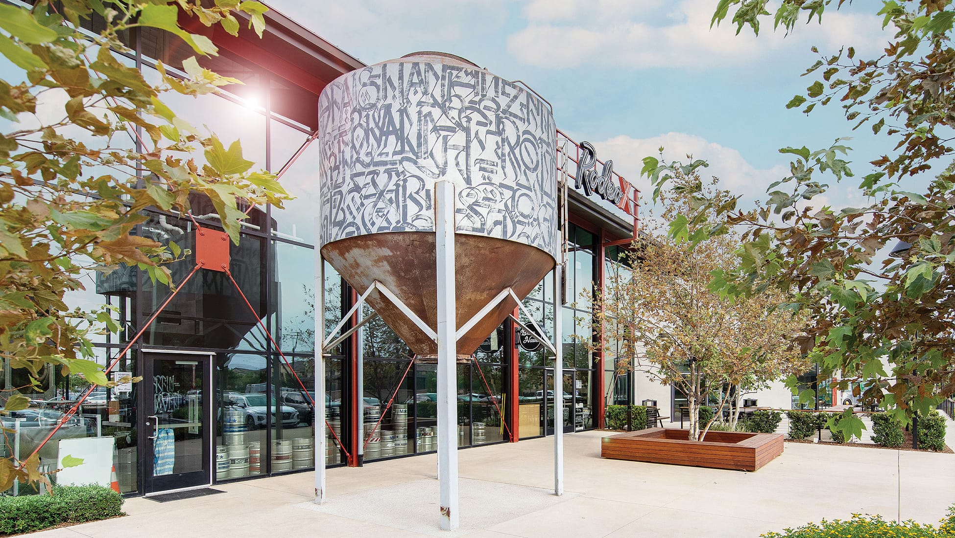 Large repurposed grain silo painted with abstract calligraphic graffiti lettering, mounted on white support beams outside a glass-walled brewery with 'Rodeo X' signage and a 'New Haven' circular logo, beer kegs visible through the windows and a wood planter with a tree nearby.