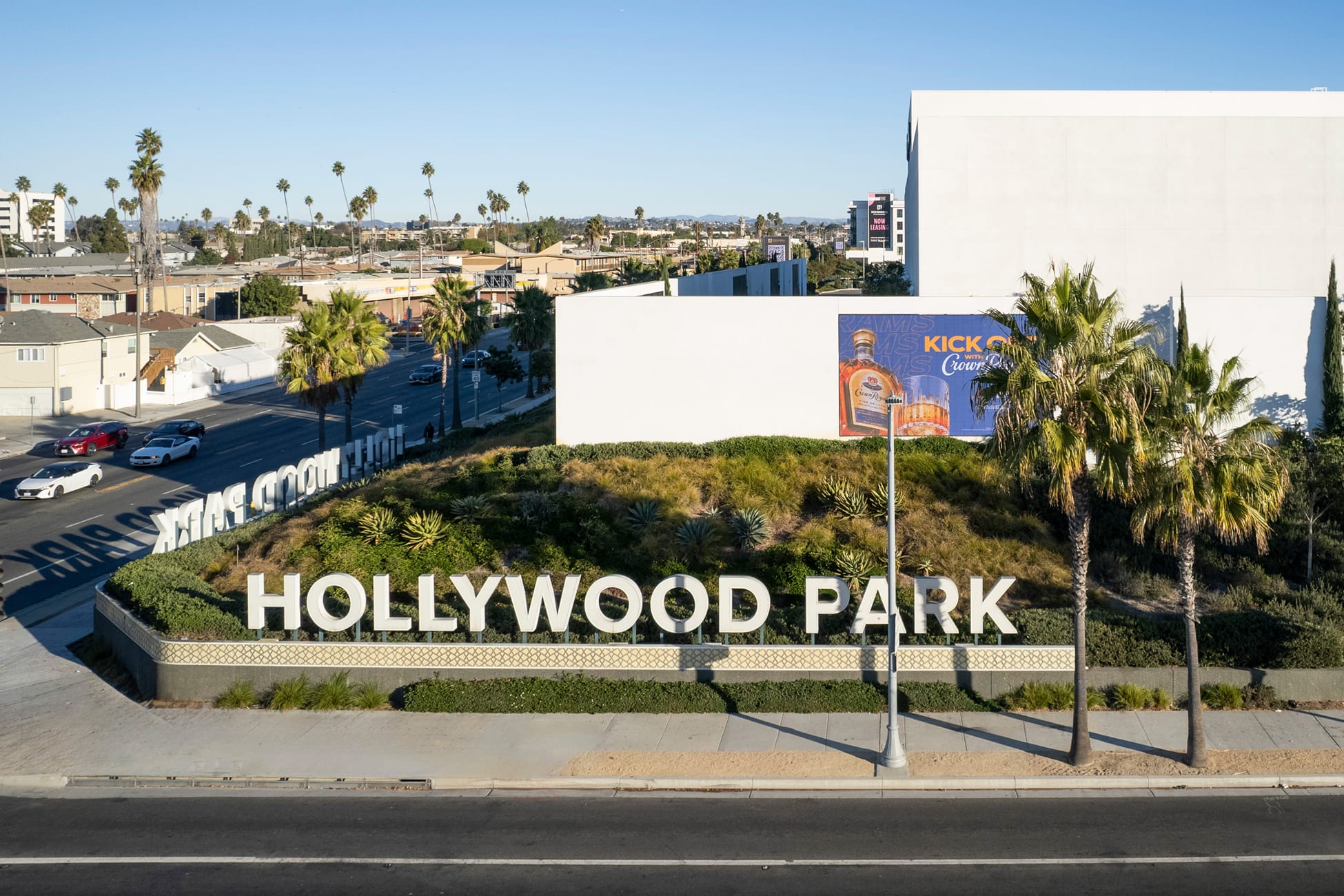 Aerial view of Hollywood Park large-scale white dimensional letter monument sign on landscaped berm with drought-tolerant plants, palm trees, and Inglewood California cityscape in background
