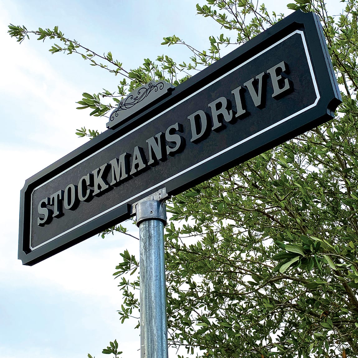Stockmans Drive street sign near Hotel Drover in the Fort Worth Stockyards, Texas, featuring decorative scroll detail and classic serif typography against a blue sky and green tree canopy