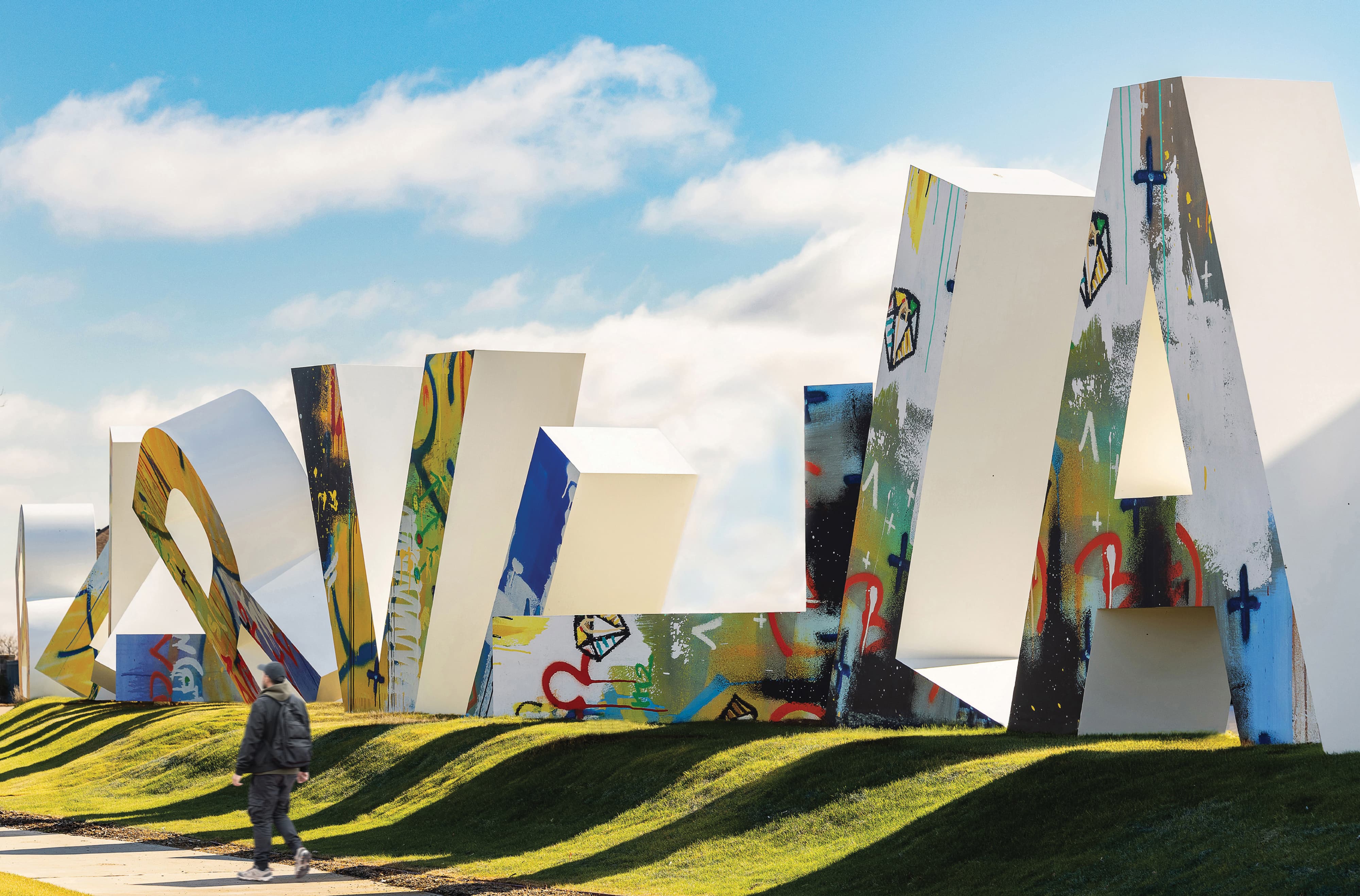 Seen from mid-distance, a person walks on a sidewalk. Behind them stands a line of large sculptures, which tower over them in height. The letters are variously rotated, and don't spell any recognizable words in this photo. The front of each letter is painted in a street art style.