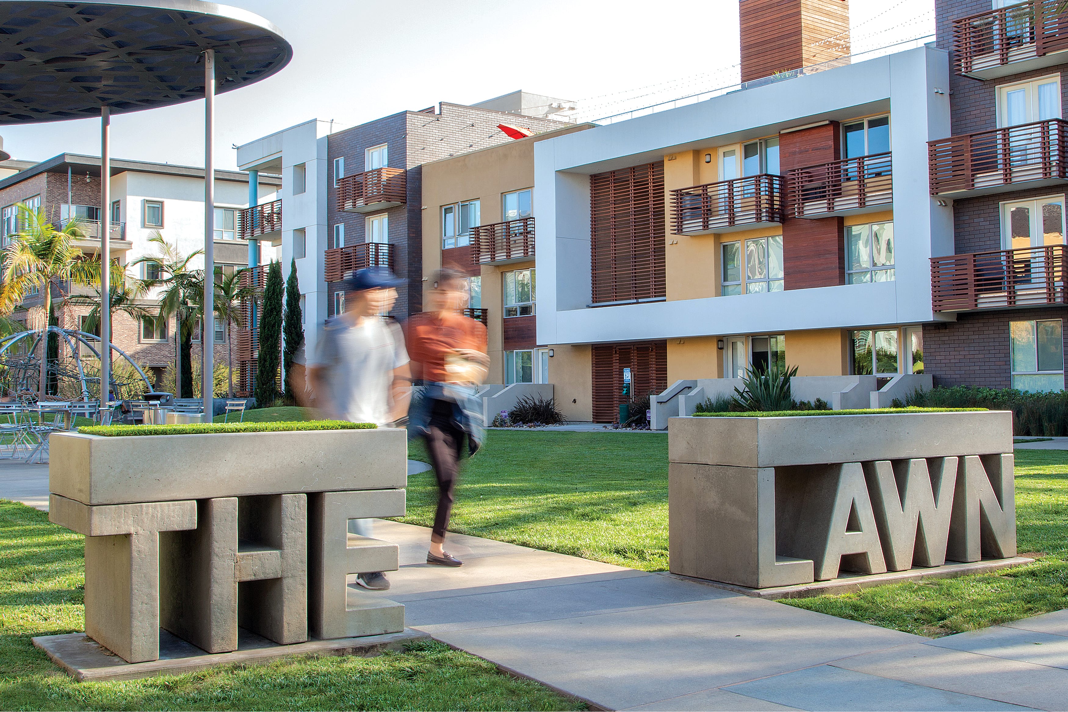 Concrete block letters spelling "THE LAWN," designed by RSM Design, doubling as planters and seating on a grassy park area at Playa Vista, with residential buildings in the background.