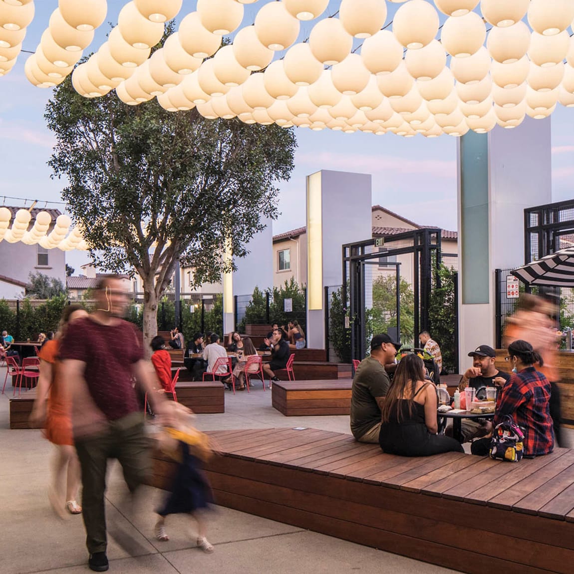 Lively outdoor courtyard at Rodeo 39 Public Market at dusk, with guests dining and gathering on tiered wood platform seating beneath a canopy of glowing globe pendant lights, surrounded by mature trees, red café chairs, and lush greenery.